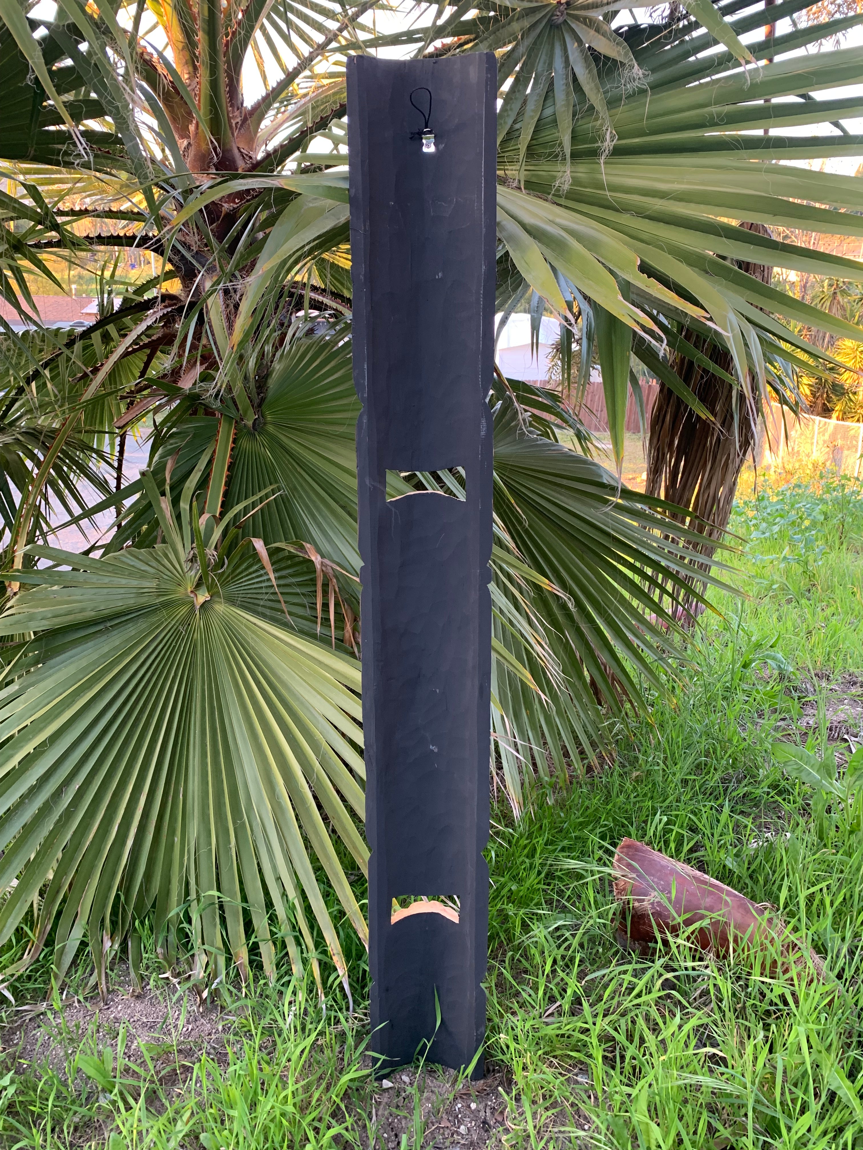 Tall dark wooden post with rectangular and arched cutouts in a garden with palm leaves