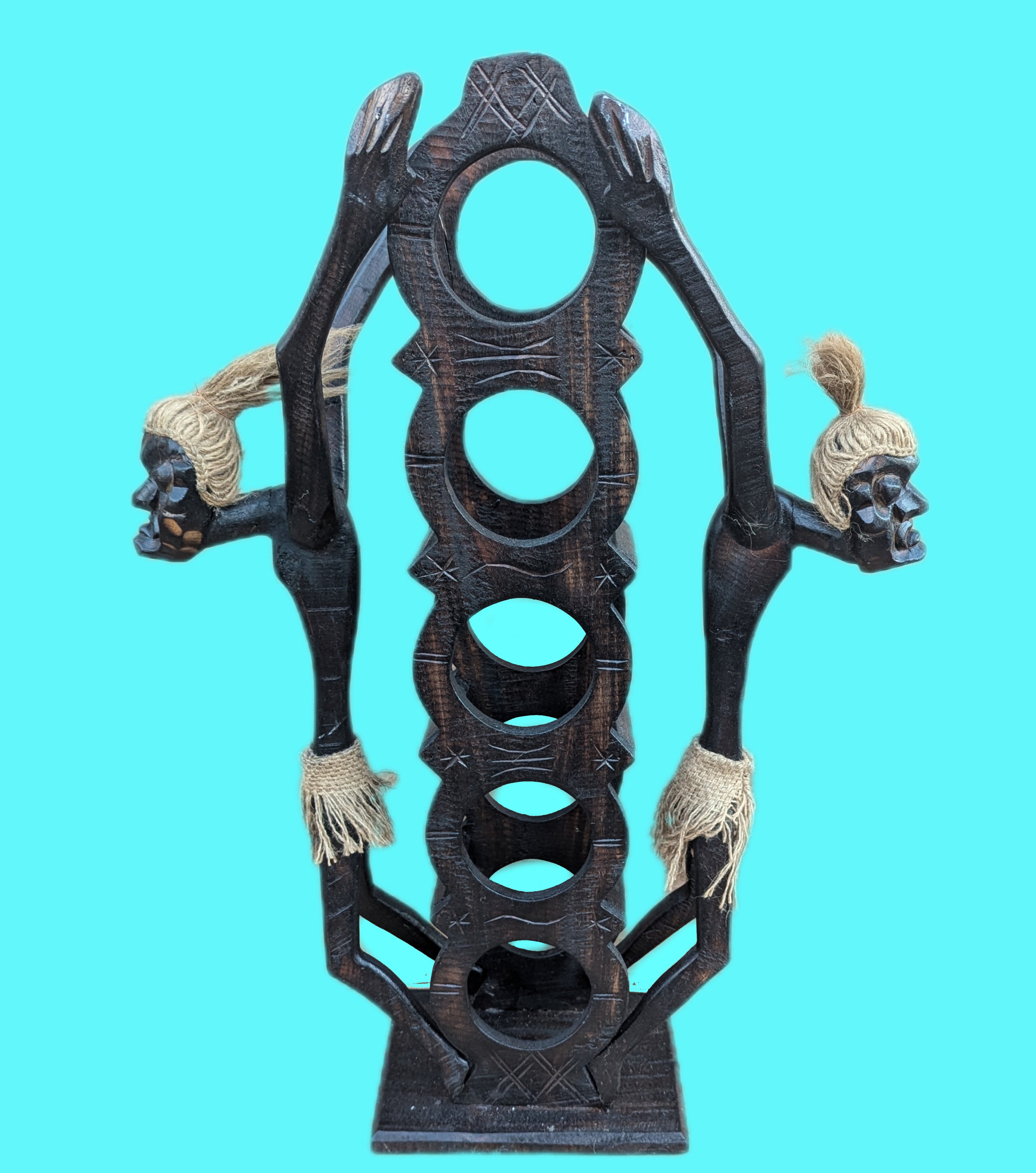Dark wooden tribal sculpture with multiple circular openings and two rope-wrapped skull figures.