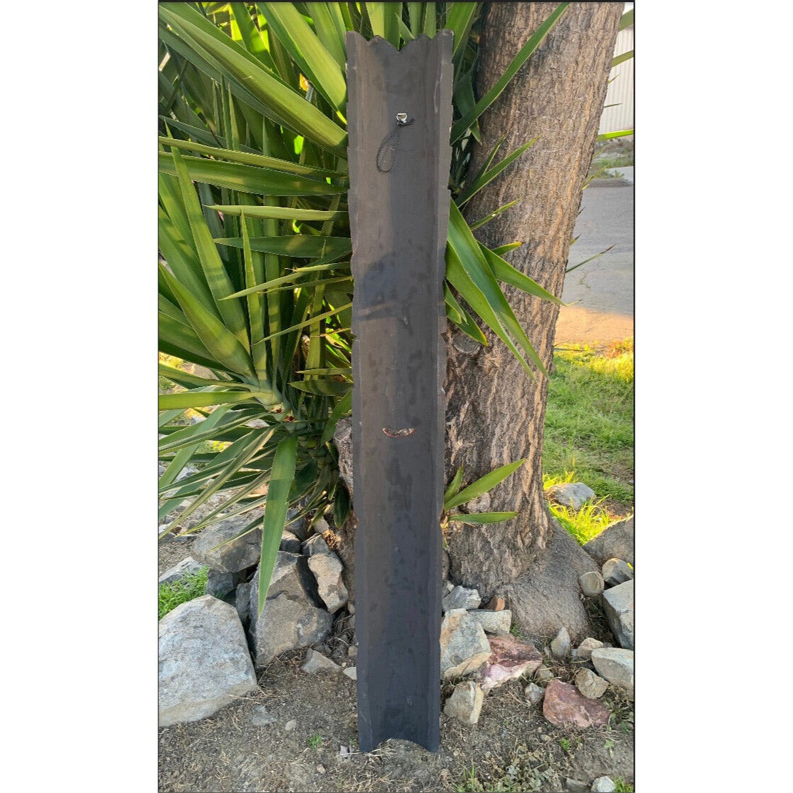 Dark vertical wooden post with scalloped top in a garden beside rocks and a tree.