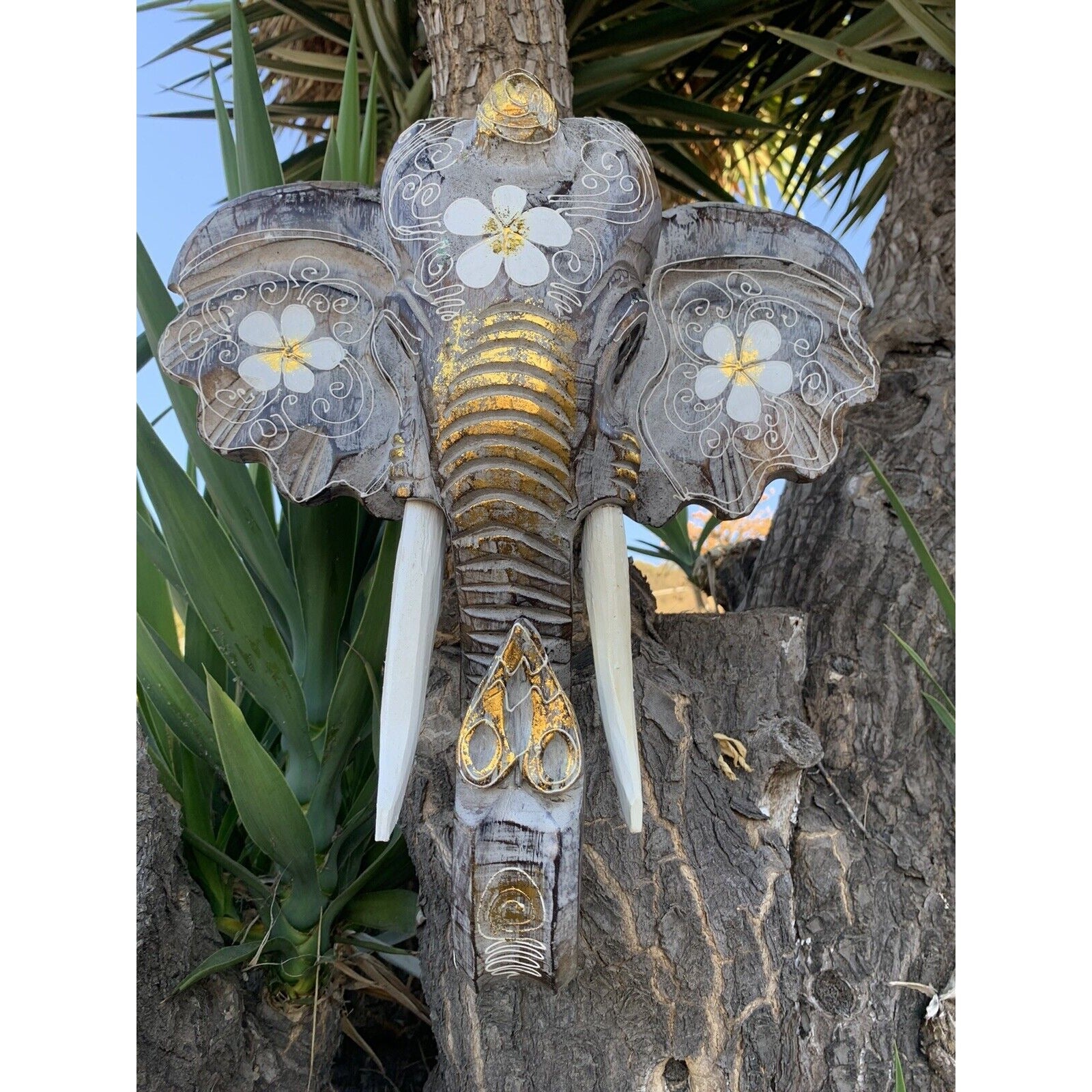 Decorative wooden elephant head sculpture with white floral patterns and gold accents on a tree trunk