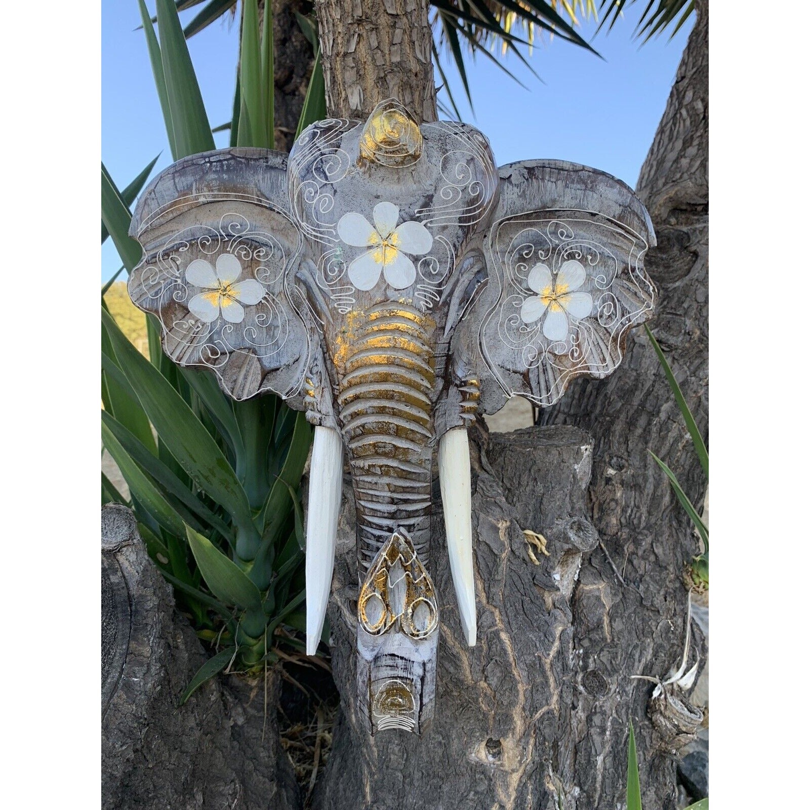 Decorative elephant head sculpture with white floral patterns and gold accents on a tree trunk