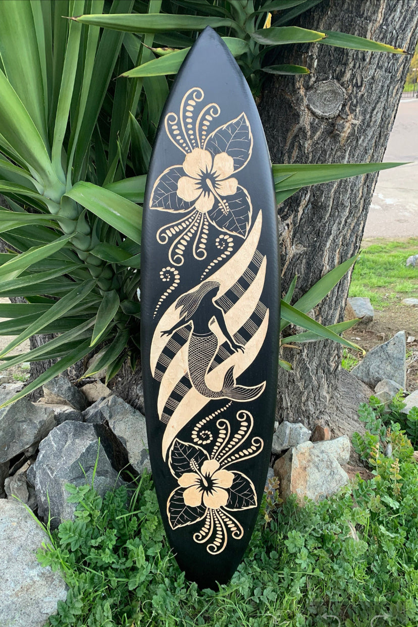 Black surfboard with beige tropical flowers, leaves, and a mermaid silhouette leaning against a tree