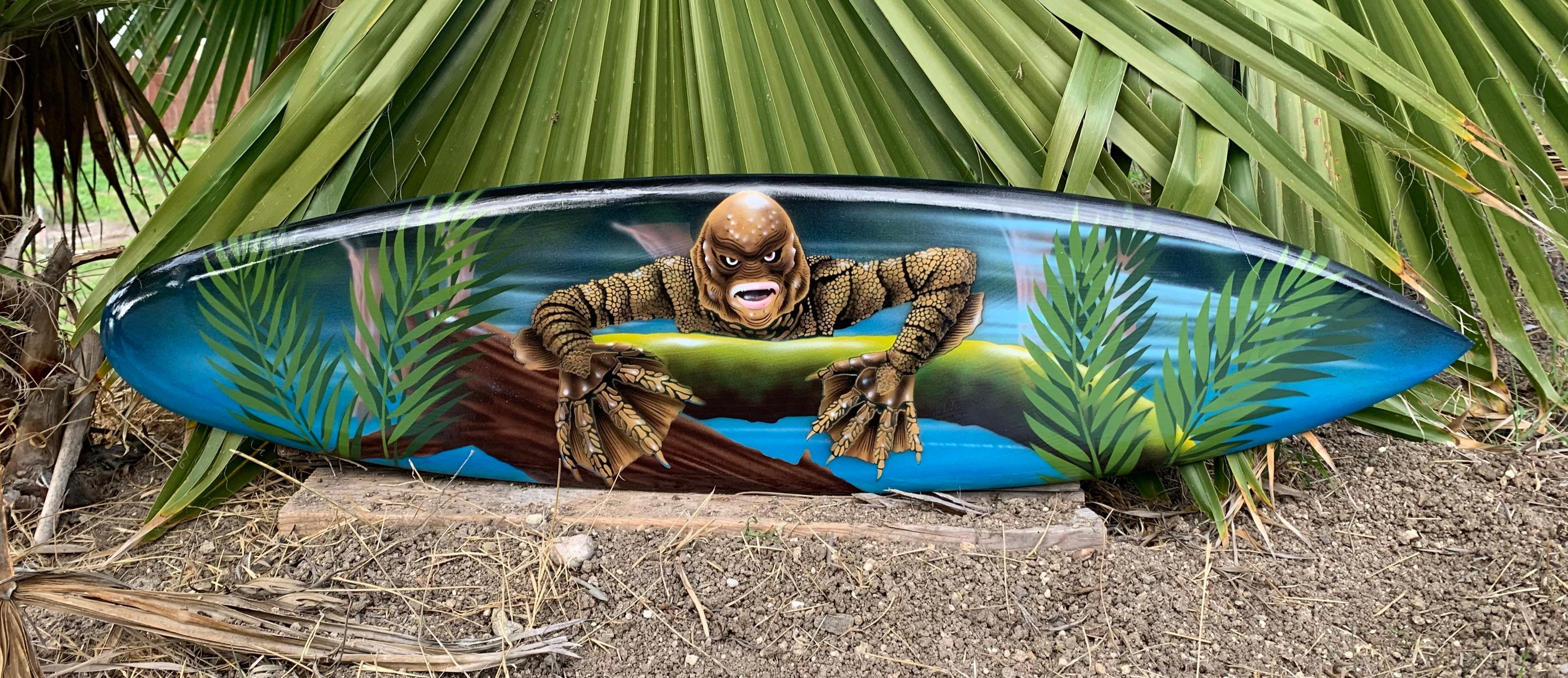decorative surfboard featuring a menacing creature painting among tropical palm leaves