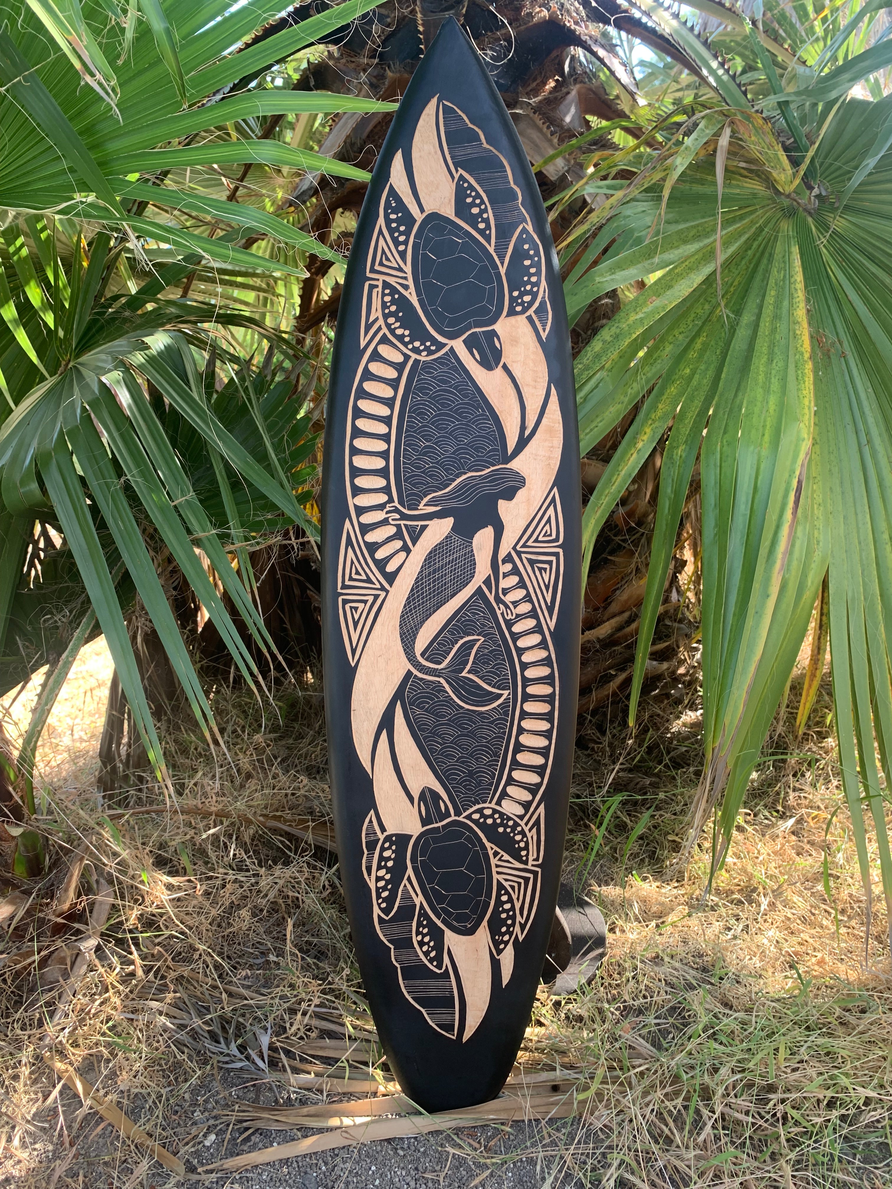 Longboard-style surfboard with tan tribal turtle design on black surface, propped among palm fronds.