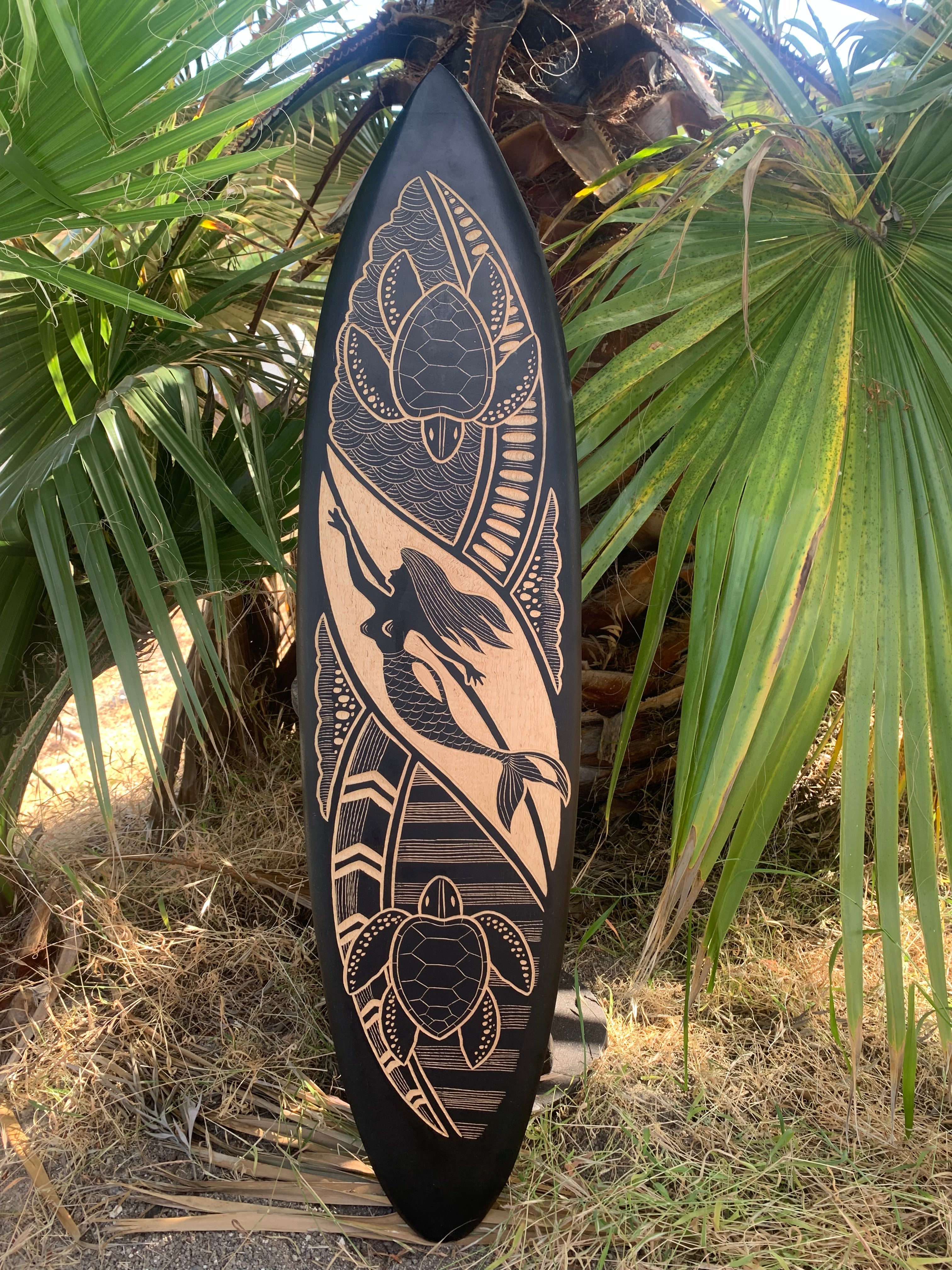 Decorative wooden surfboard featuring a mermaid and turtle motifs carved in light wood on black background