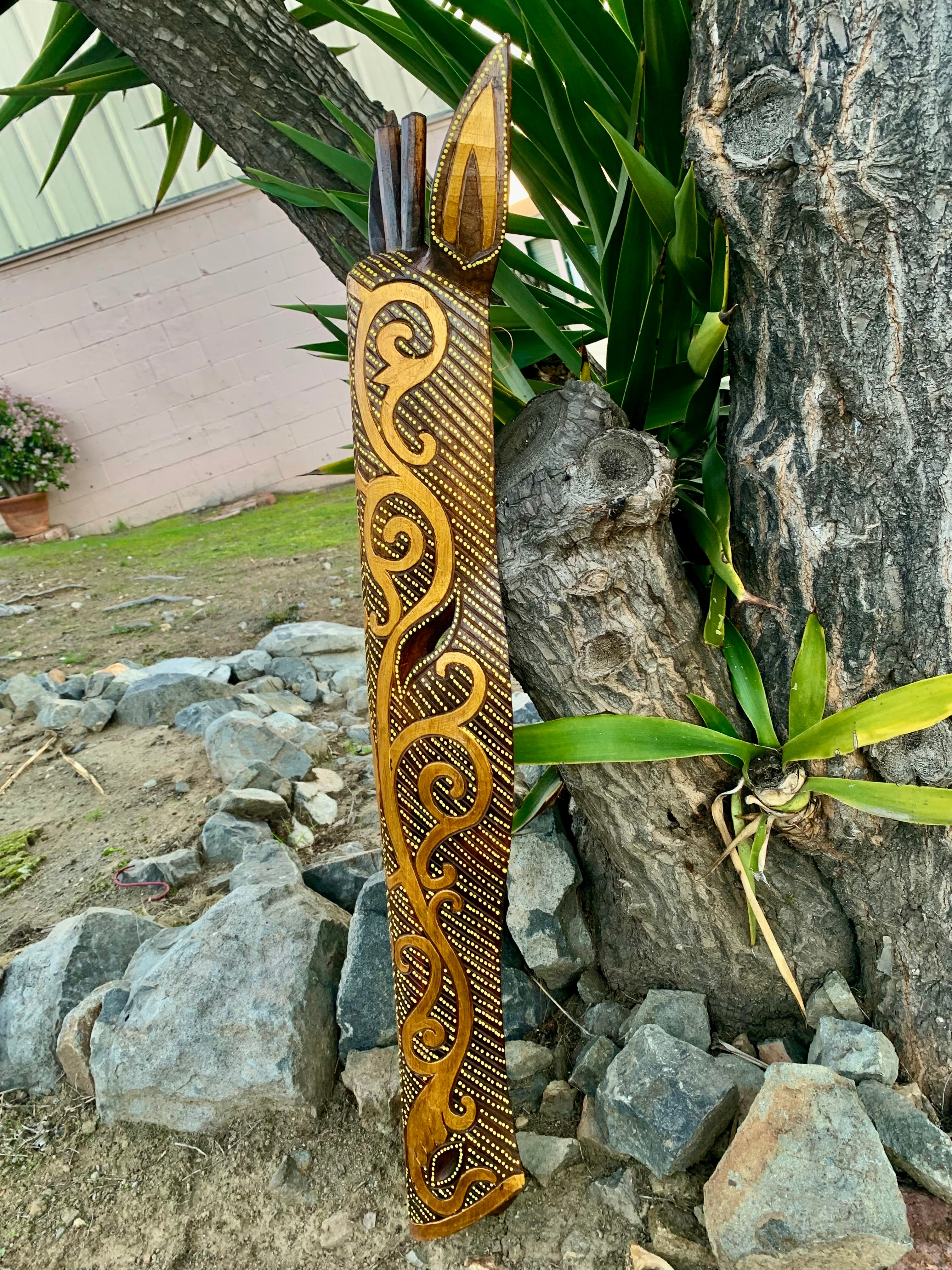 Hand-carved wooden totem pole with gold scroll patterns leaning against a tree.