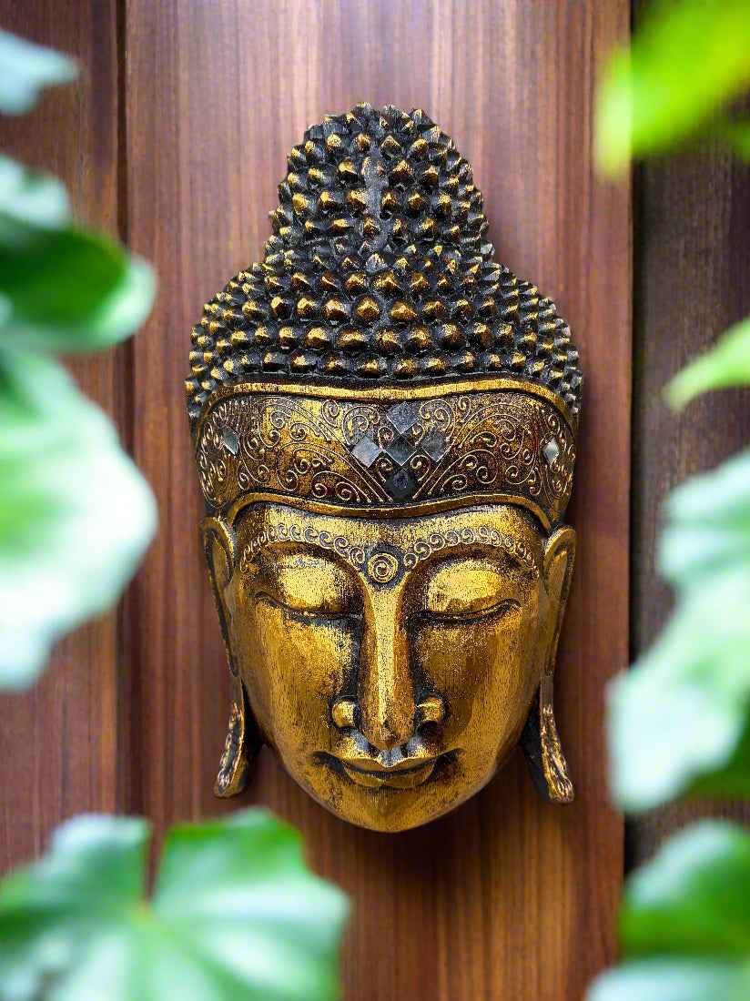 Golden Buddha head wall sculpture with ornate headdress on a wooden panel, framed by greenery.