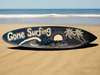 Surfboard with 'Gone Surfing' graphic on a sandy beach by the ocean.