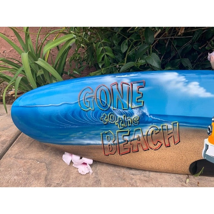 Blue surfboard with Gone to the Beach graphic on sandy ground beside green plants.