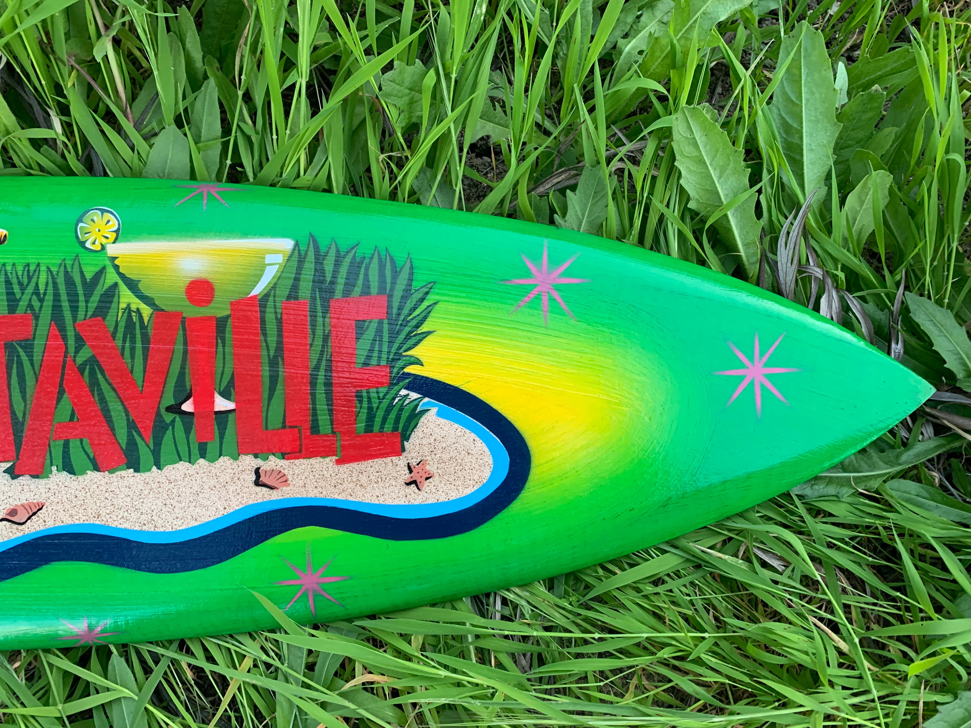 Green tropical-themed surfboard with a beach scene and pink starbursts resting in tall grass