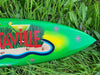 Green tropical-themed surfboard with a beach scene and pink starbursts resting in tall grass