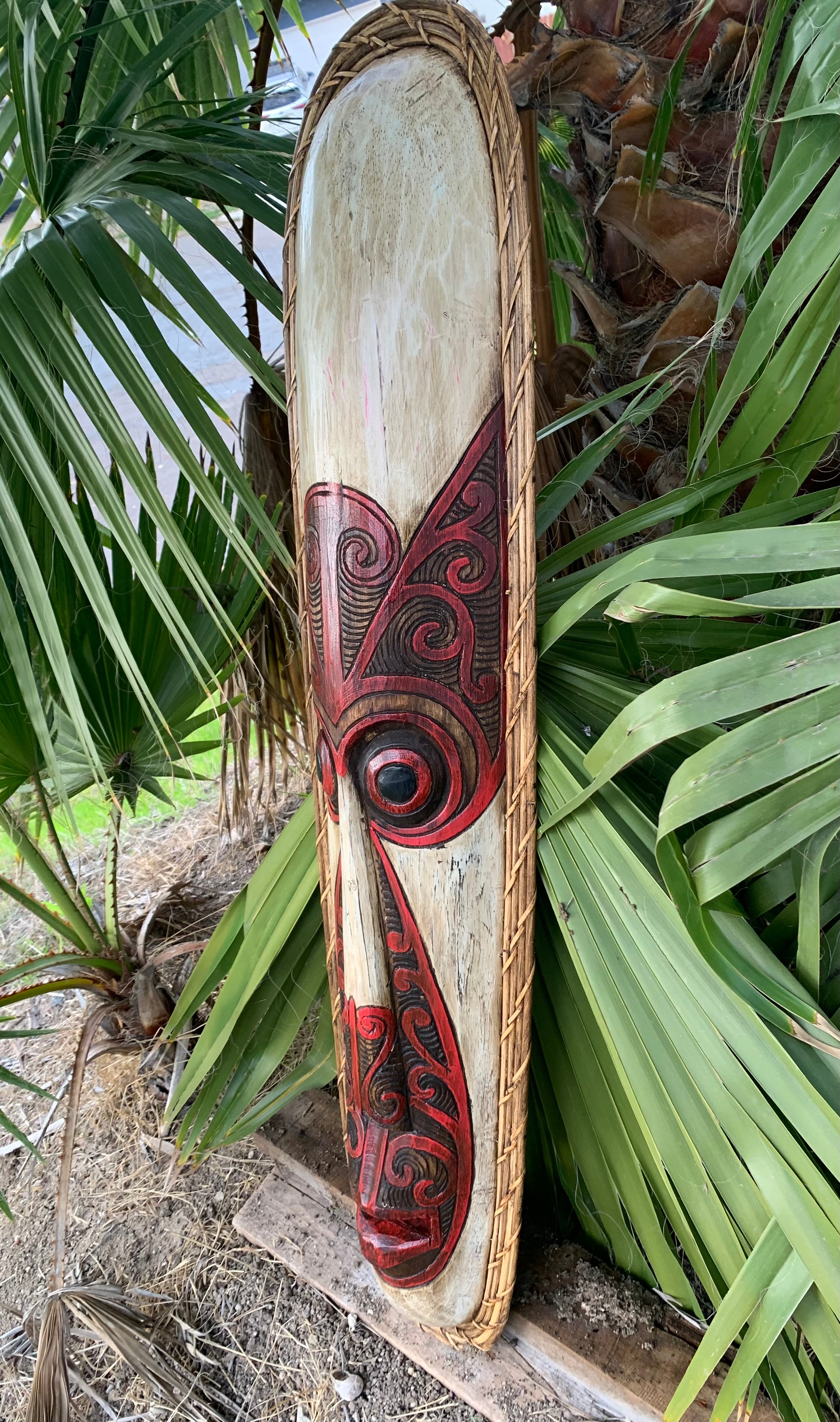 Hand-carved wooden paddle with red and black koru patterns leaning against palm leaves.