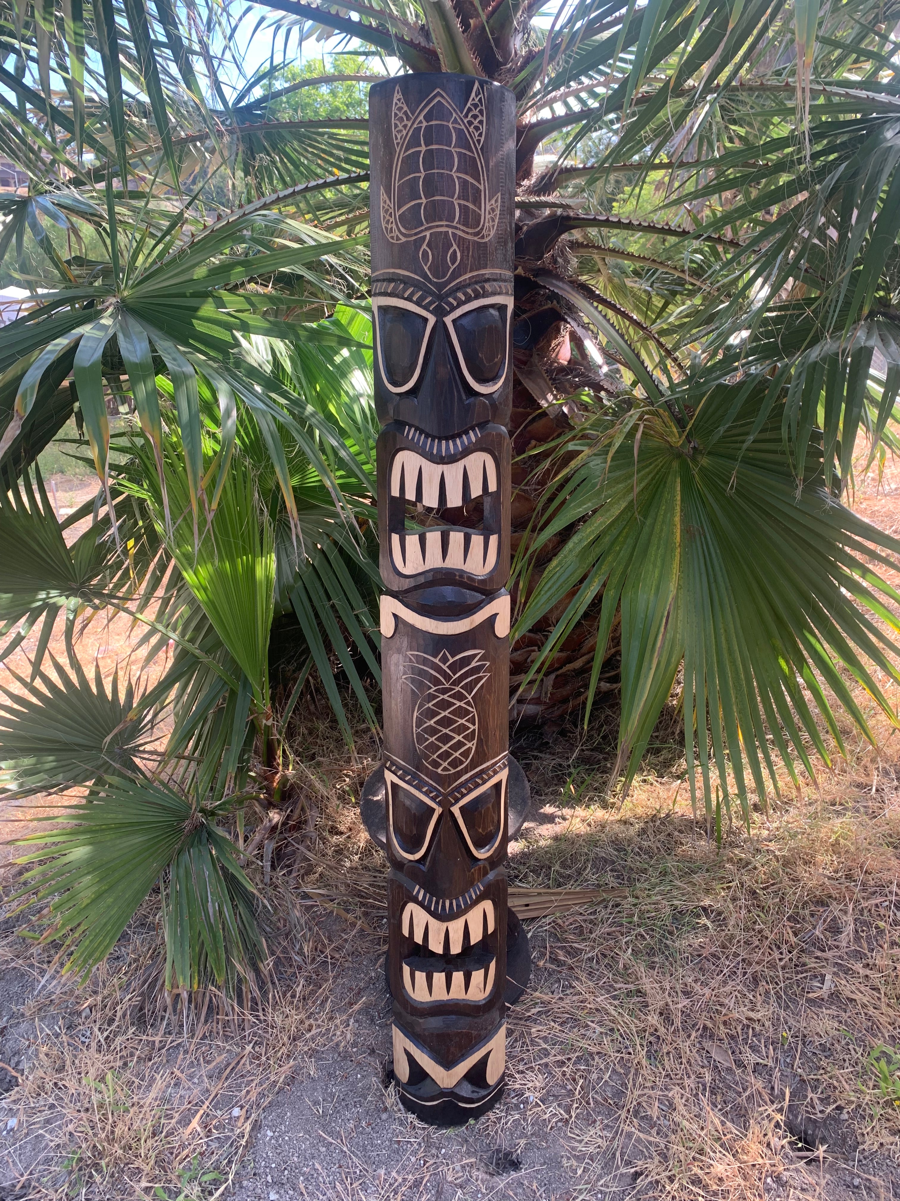 Hand-carved tiki totem pole with multiple faces beside palm leaves outdoors