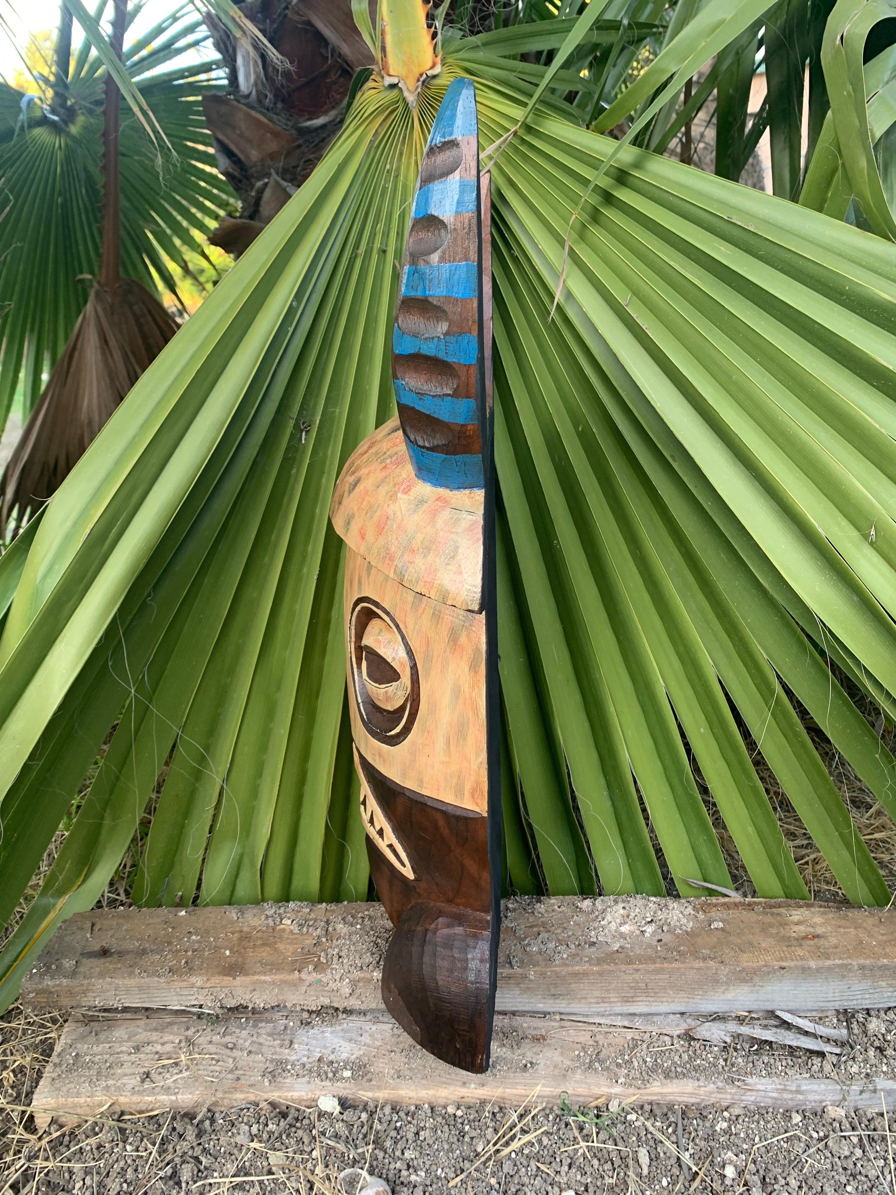 Hand-carved tiki wood sculpture with blue stripes and an eye motif beside palm leaves.