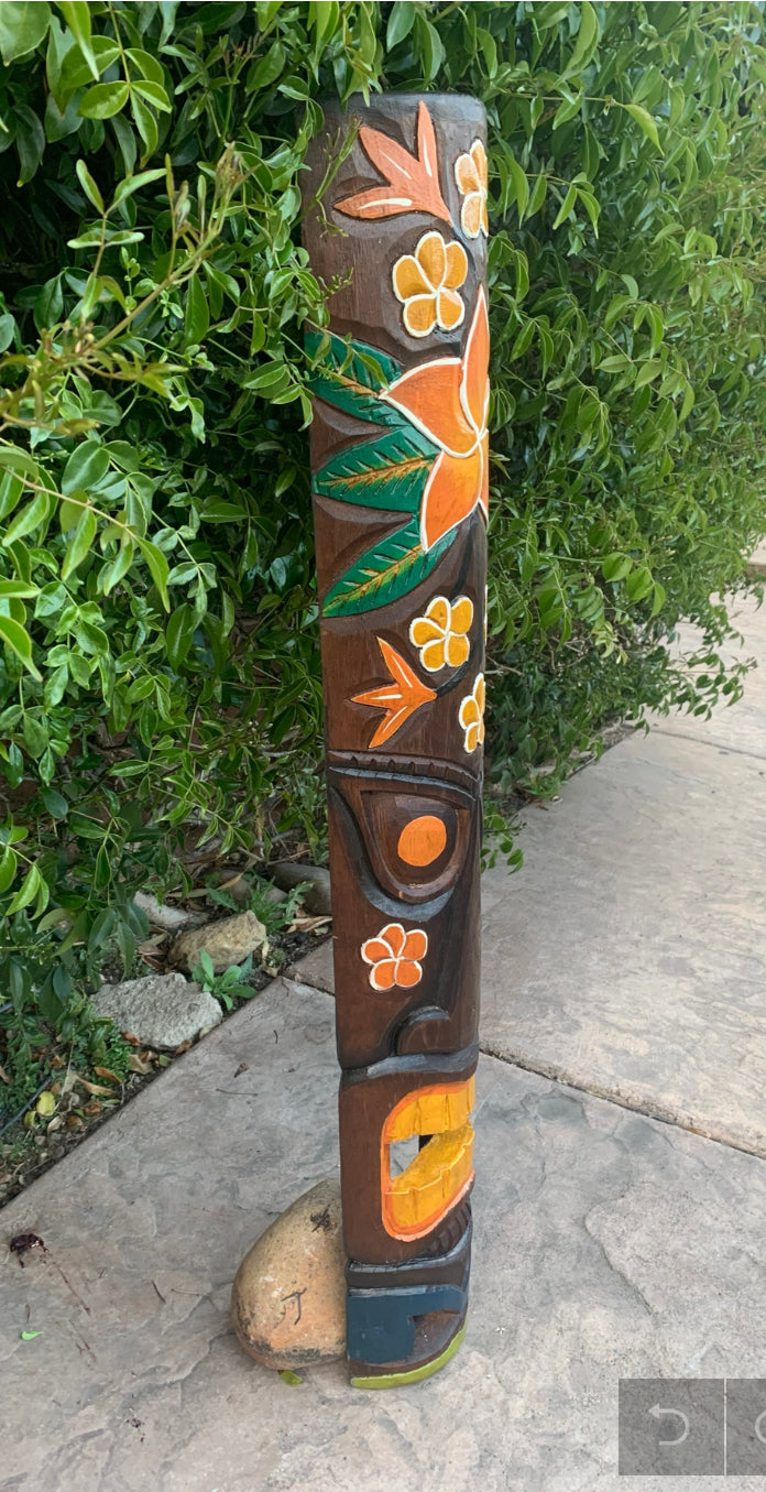 Outdoor wooden totem pole carved with orange flowers and green leaves beside a hedge