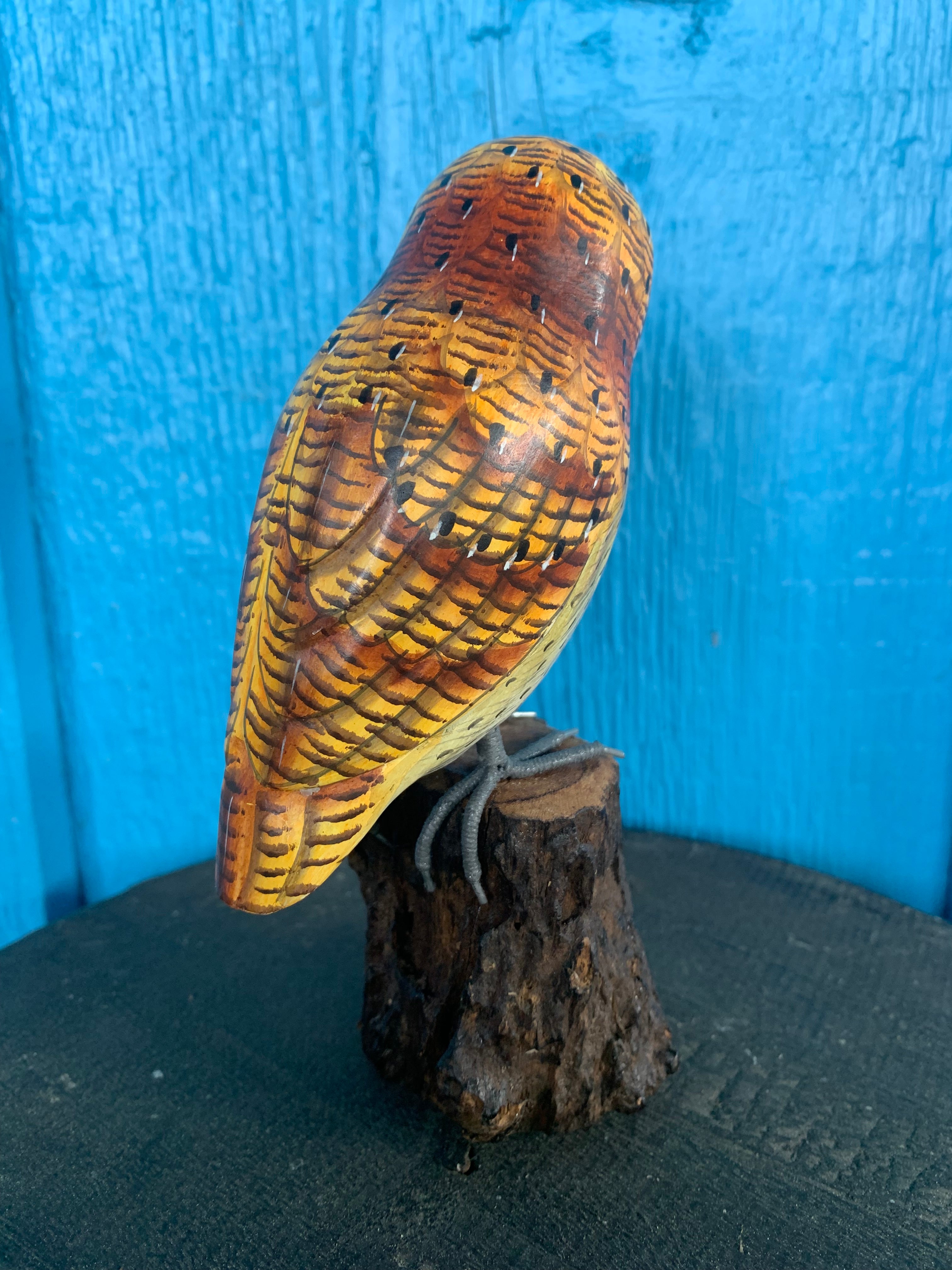 Hand-carved wooden owl sculpture with yellow-brown feathers perched on a tree stump against a blue background