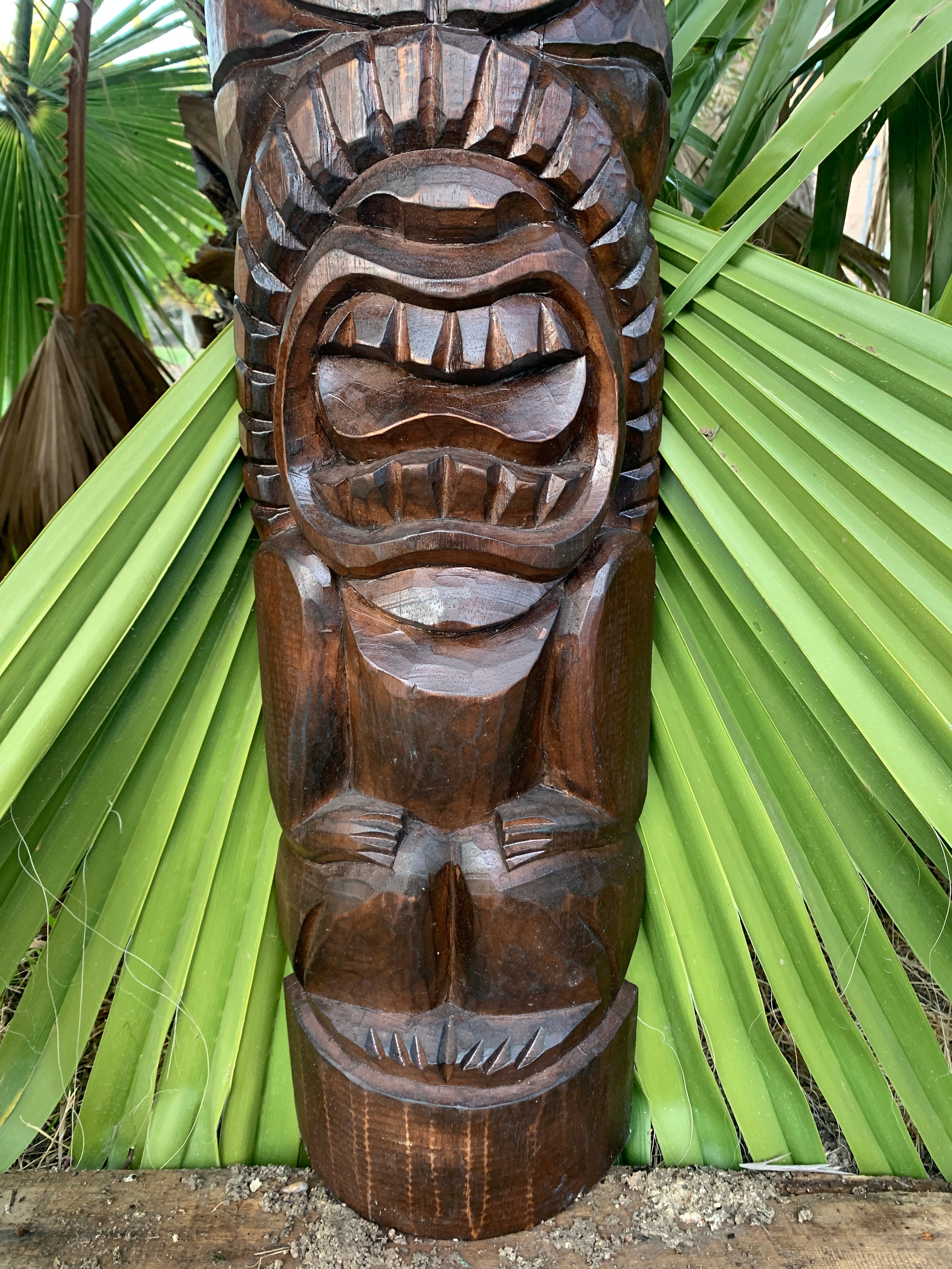 Hand-carved wooden tiki statue with a wide toothy grin among tropical palm leaves.