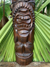 Hand-carved wooden tiki statue with a wide toothy grin among tropical palm leaves.