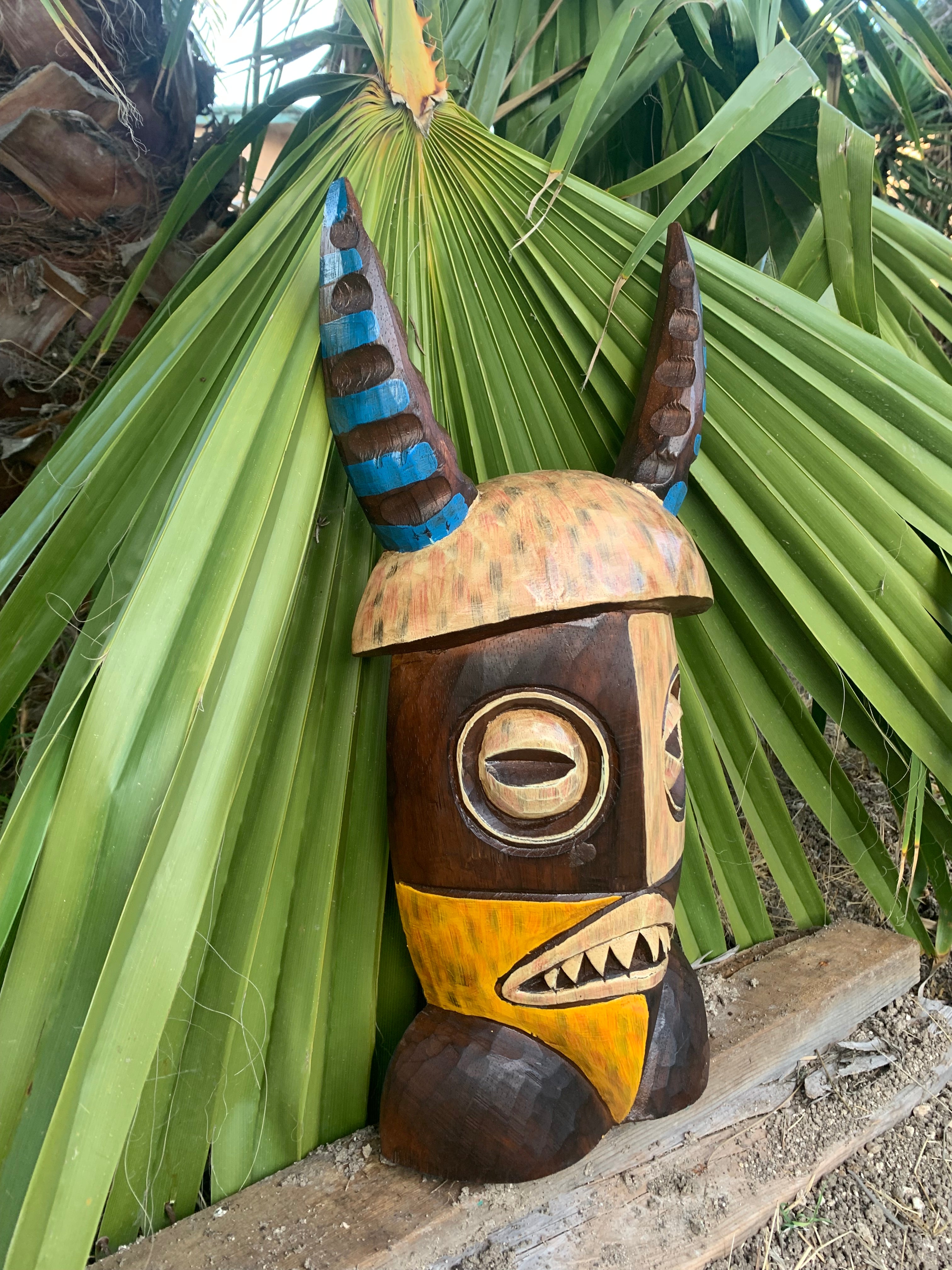 Wooden tiki statue with blue-brown horned head and yellow beak among tropical palm leaves.