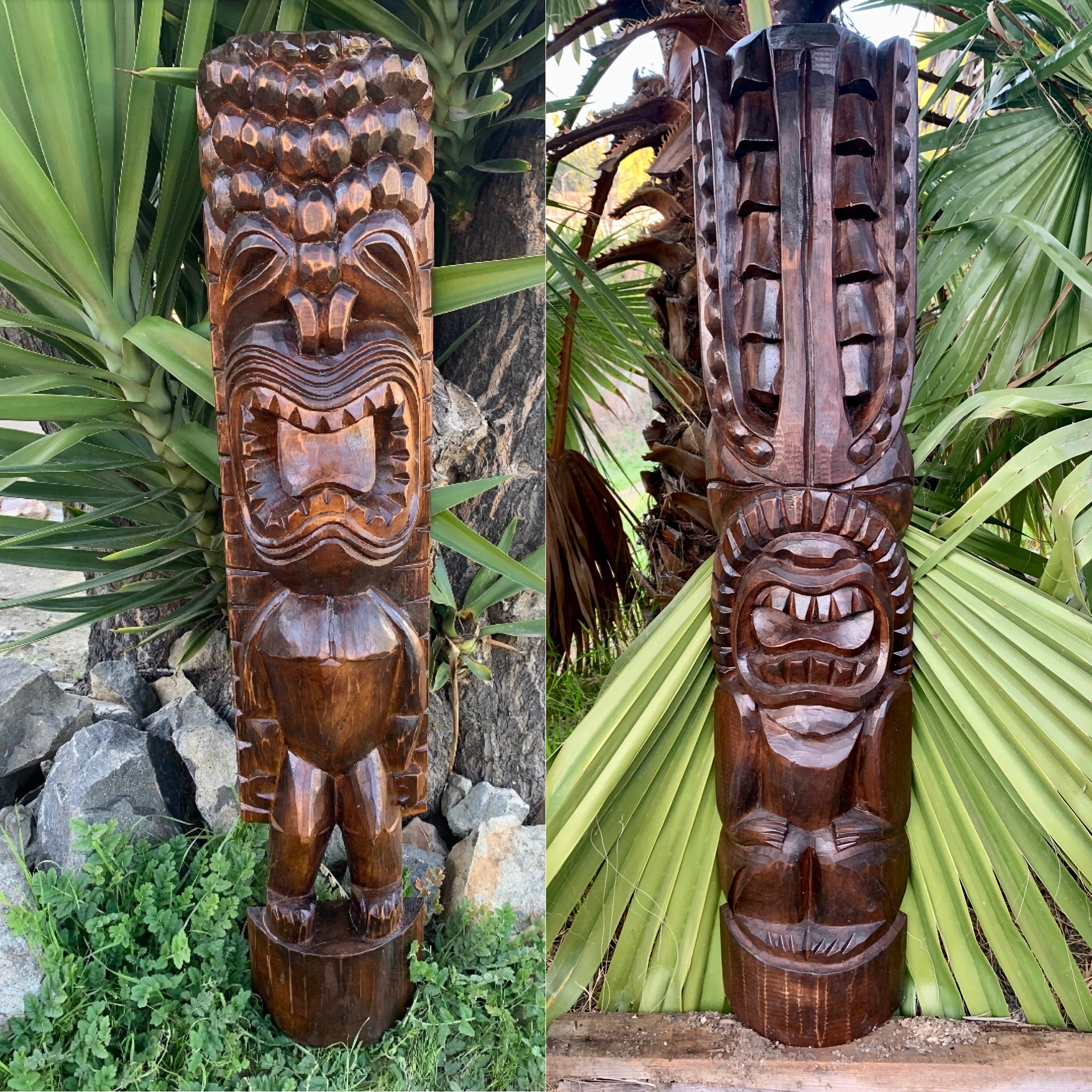 Two tall carved wooden tiki statues with fierce faces in a tropical garden.