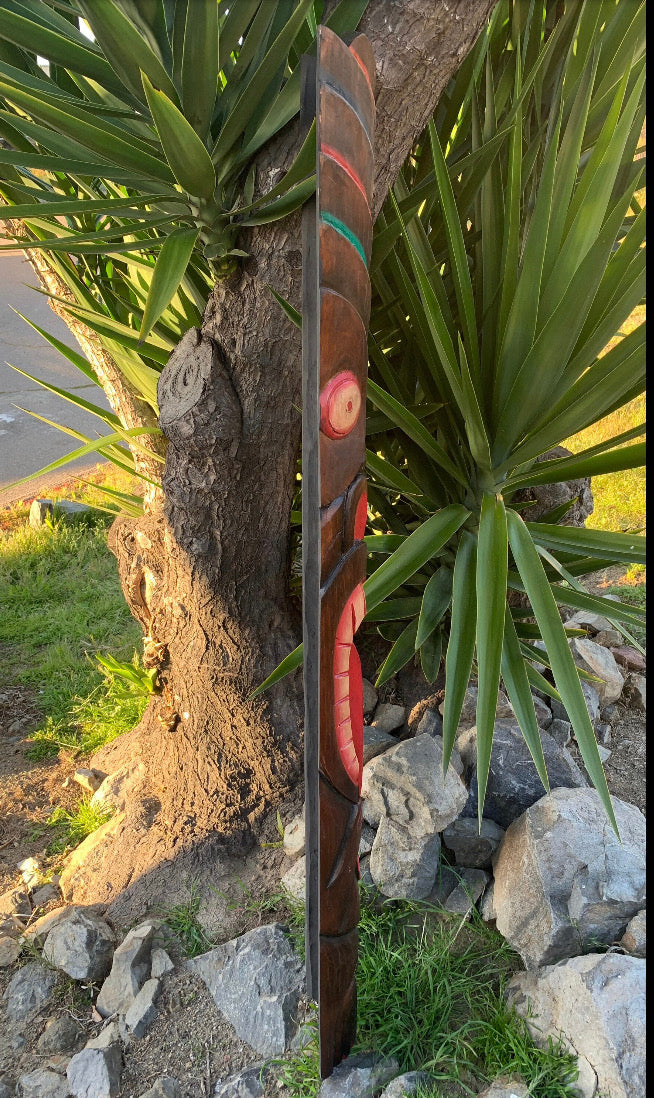 Hand-carved wooden totem pole leaning against a tree in a garden with spiky green plants and rocks