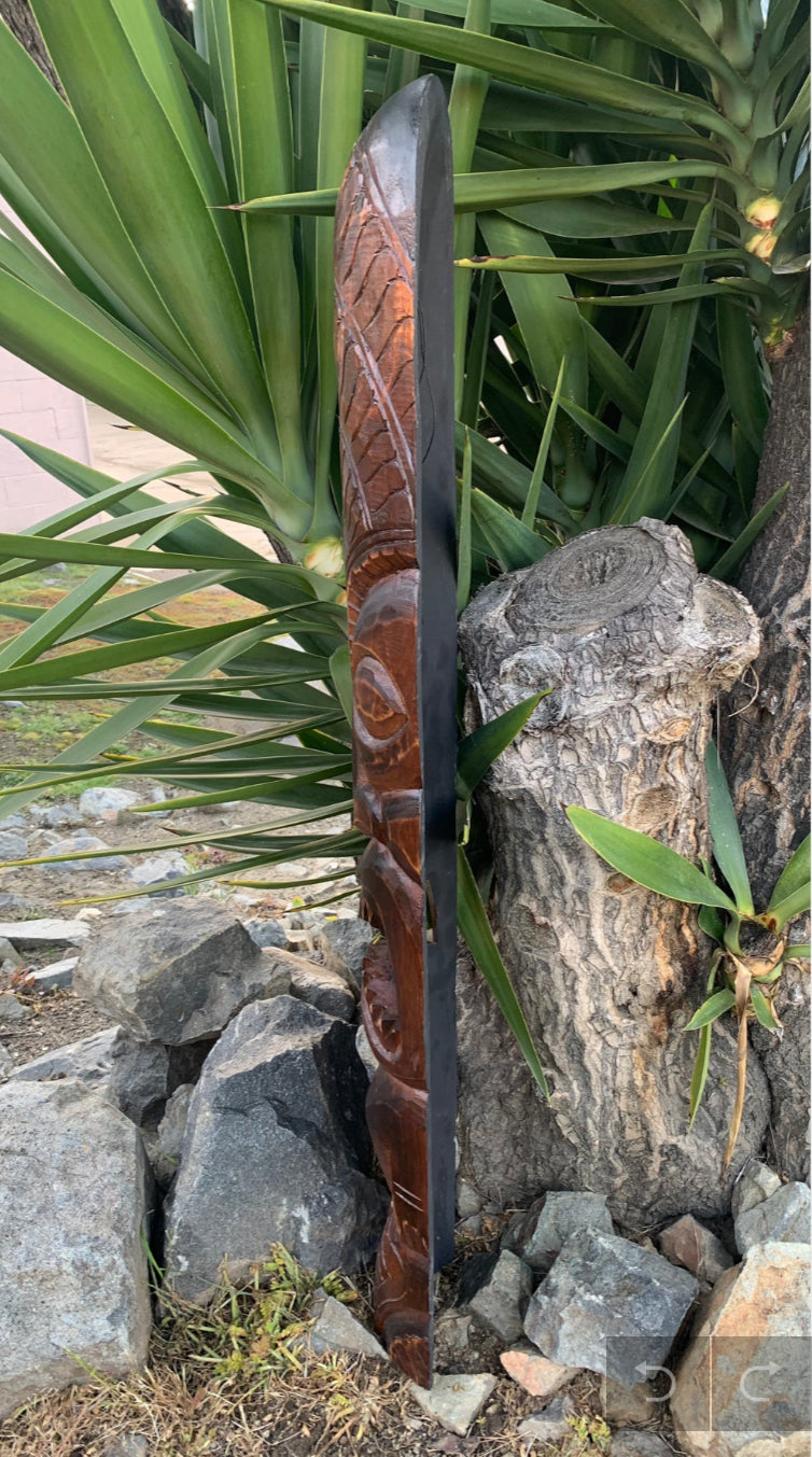 hand-carved wooden totem pole leaning against rocks with green plants in background
