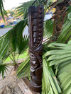 Dark wooden carved totem post with geometric patterns among palm fronds in a garden