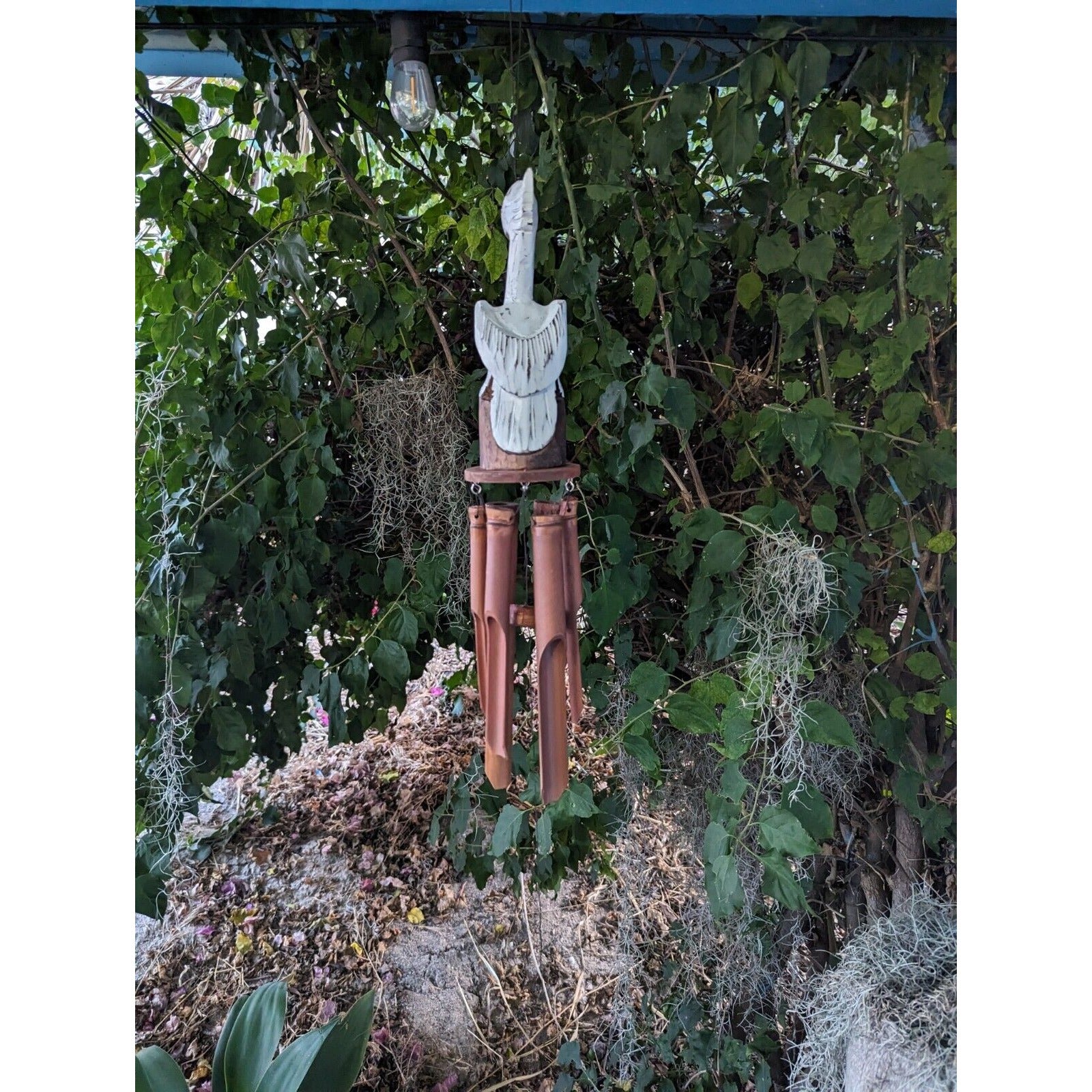 Hand-carved wooden wind chime with bamboo tubes, hanging from a carved top figure among greenery.