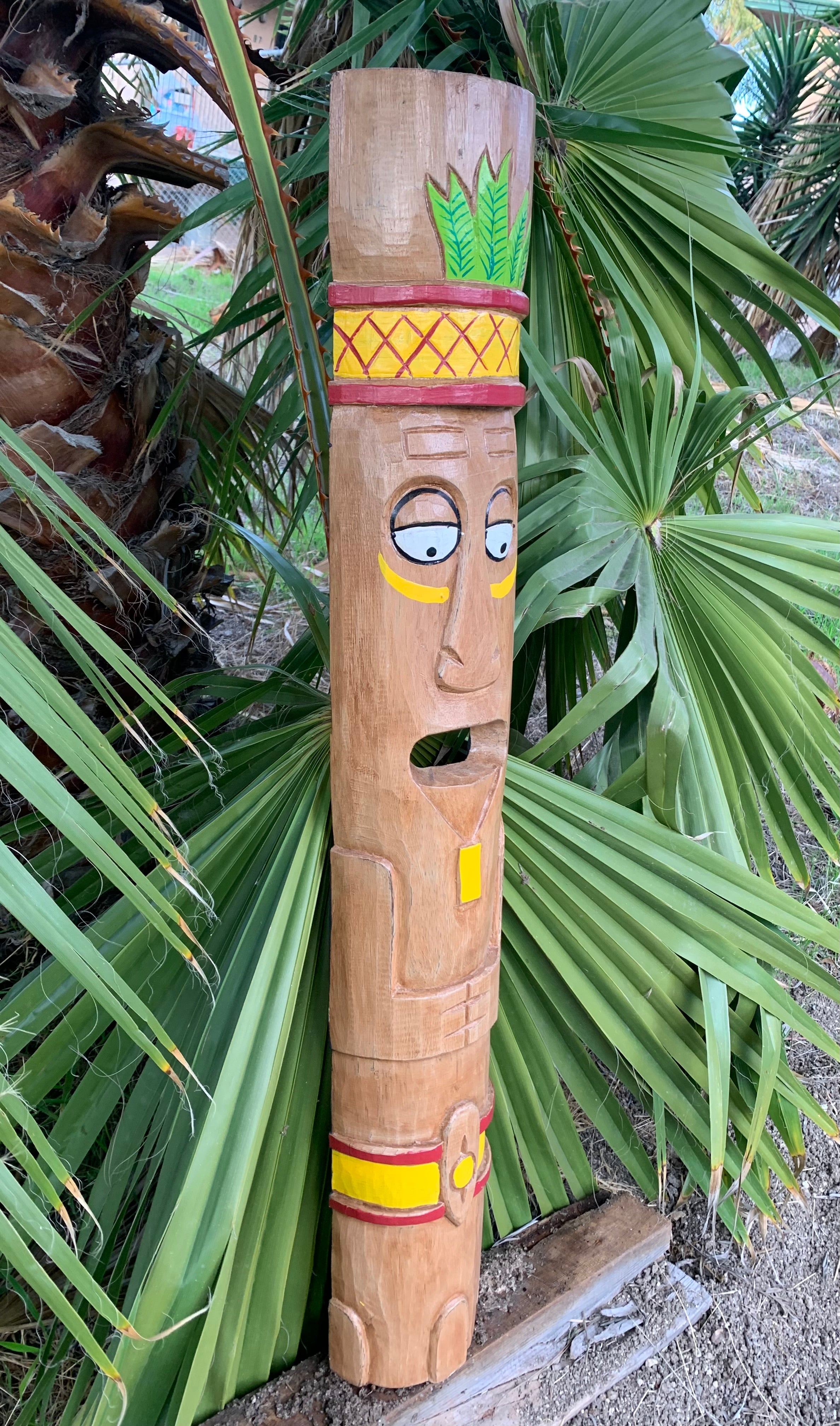 Wooden carved totem pole with a face and colorful headdress among palm leaves