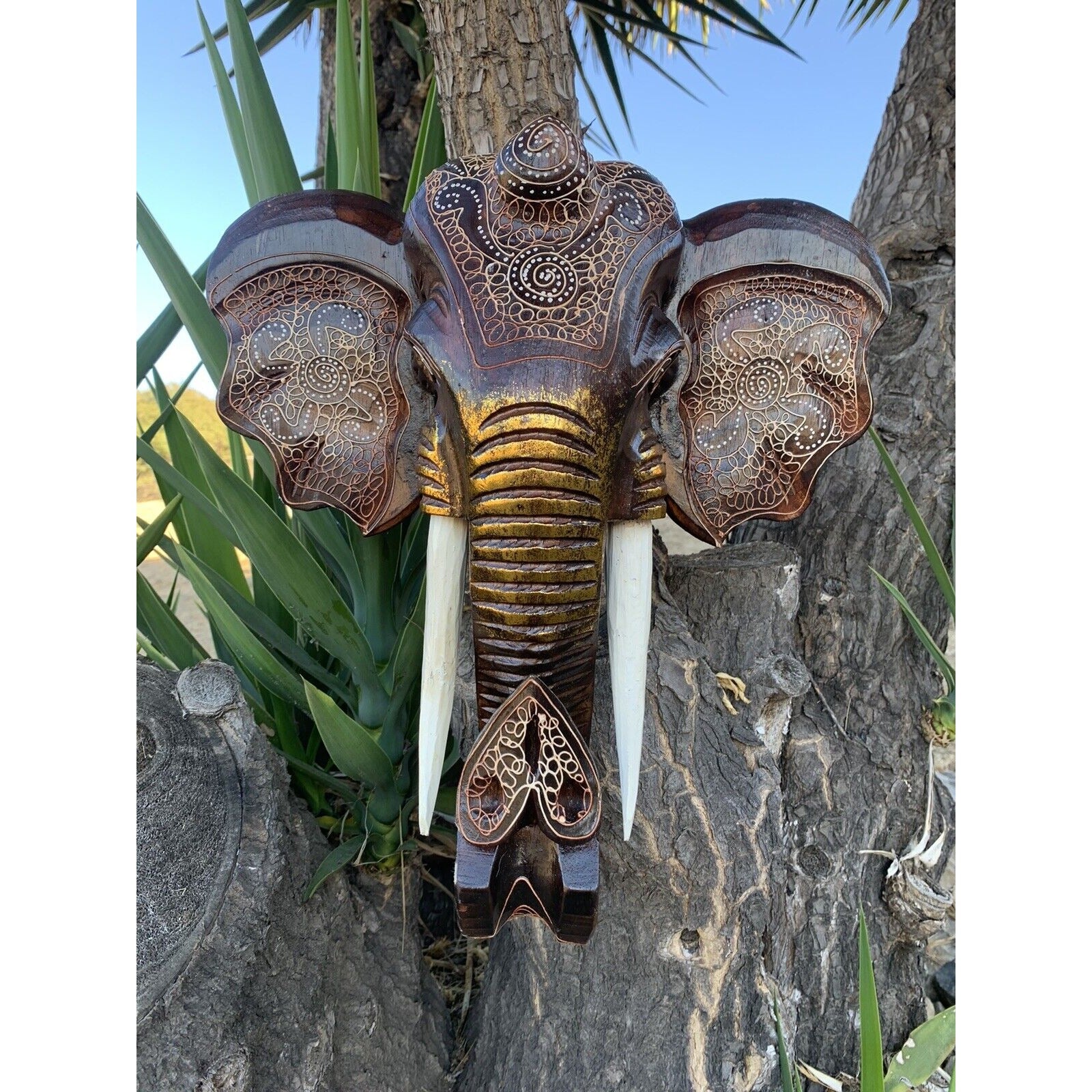 Carved wooden elephant head with ornate patterns and long tusks mounted on a tree.
