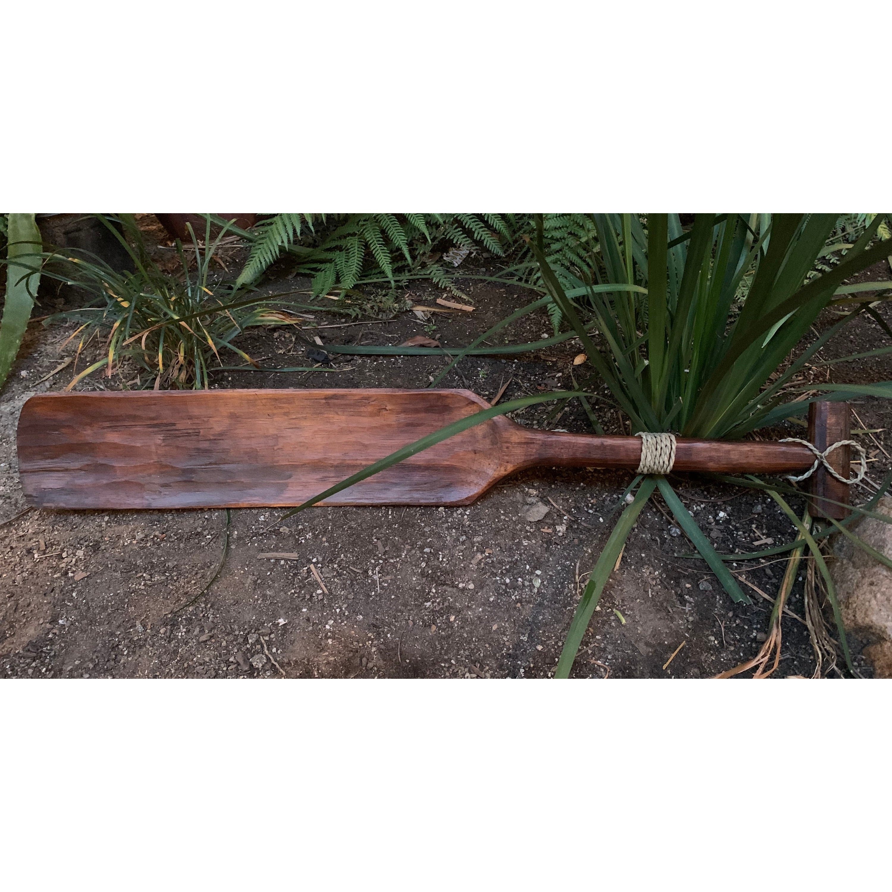 Wooden oar with rope-wrapped handle resting on dirt beside garden plants