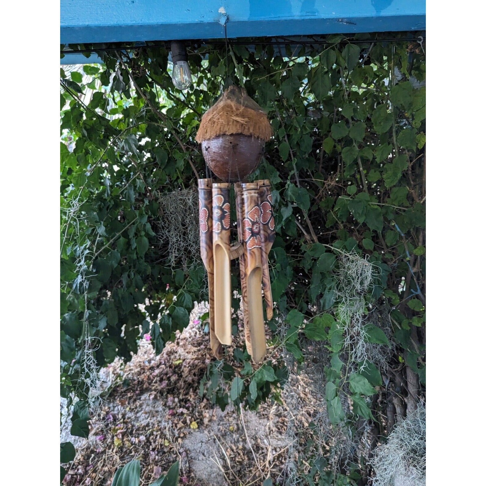 Hanging wind chime with bamboo tubes and a coconut-husk top, outdoors among green vines.