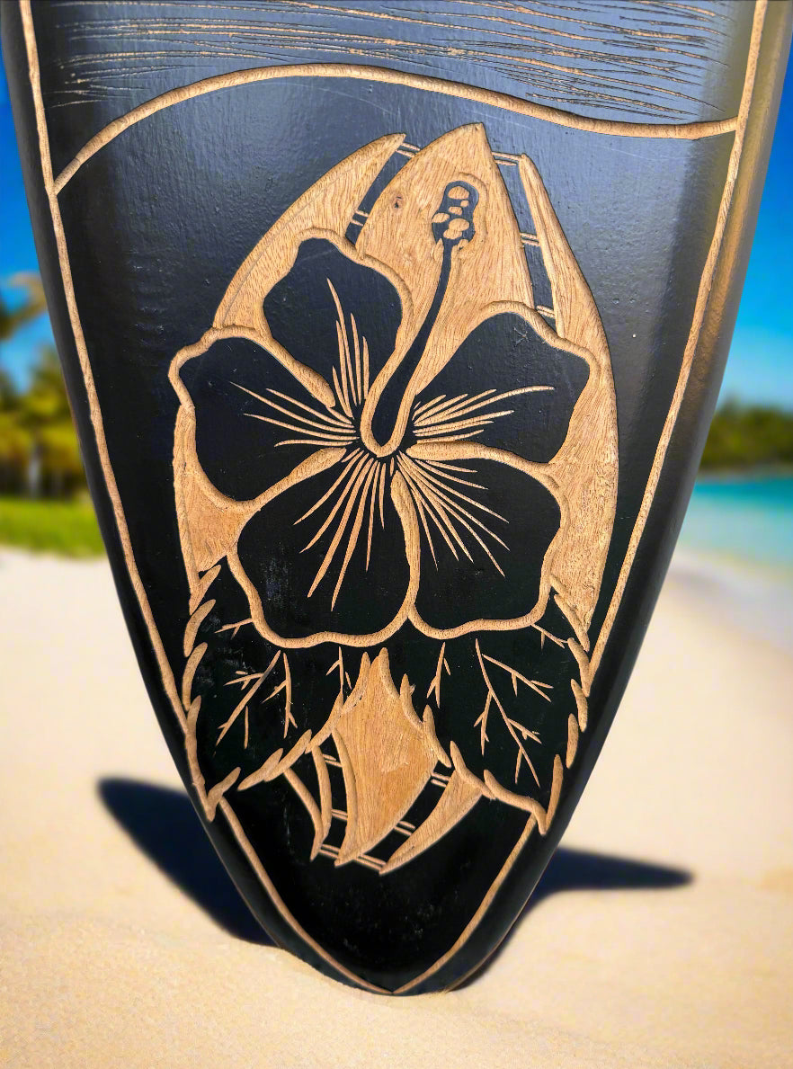 Close-up of a beige hibiscus engraving on a black surfboard with a sandy beach background