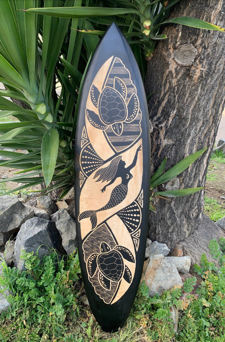 Long black surfboard with gold-tan tiki art featuring a hummingbird, turtle motifs, and flowers.