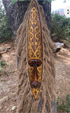Intricately carved wooden mask with gold ornate patterns and long fiber fringe hanging on a tree