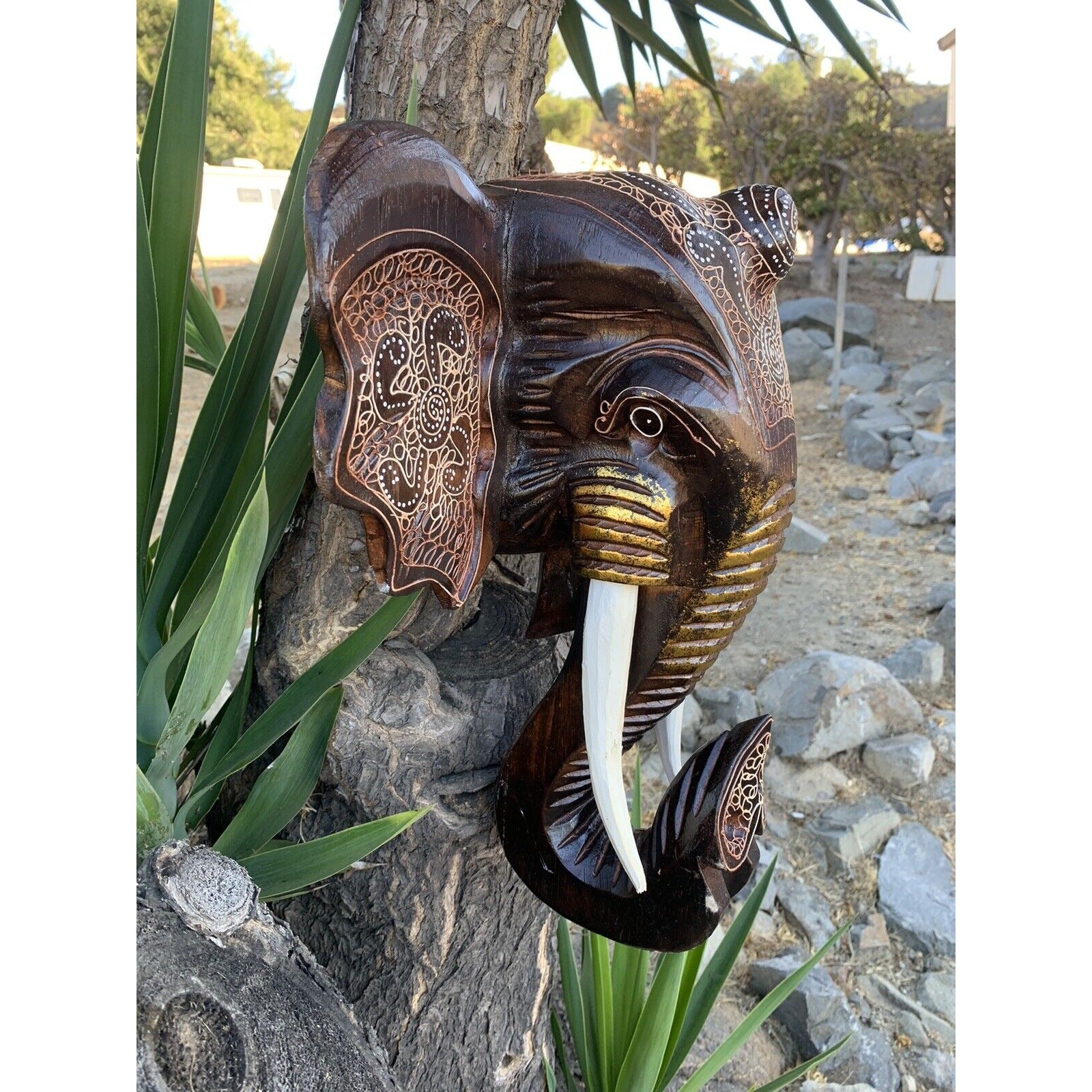 intricately carved wooden elephant head sculpture with white tusks mounted on a tree.