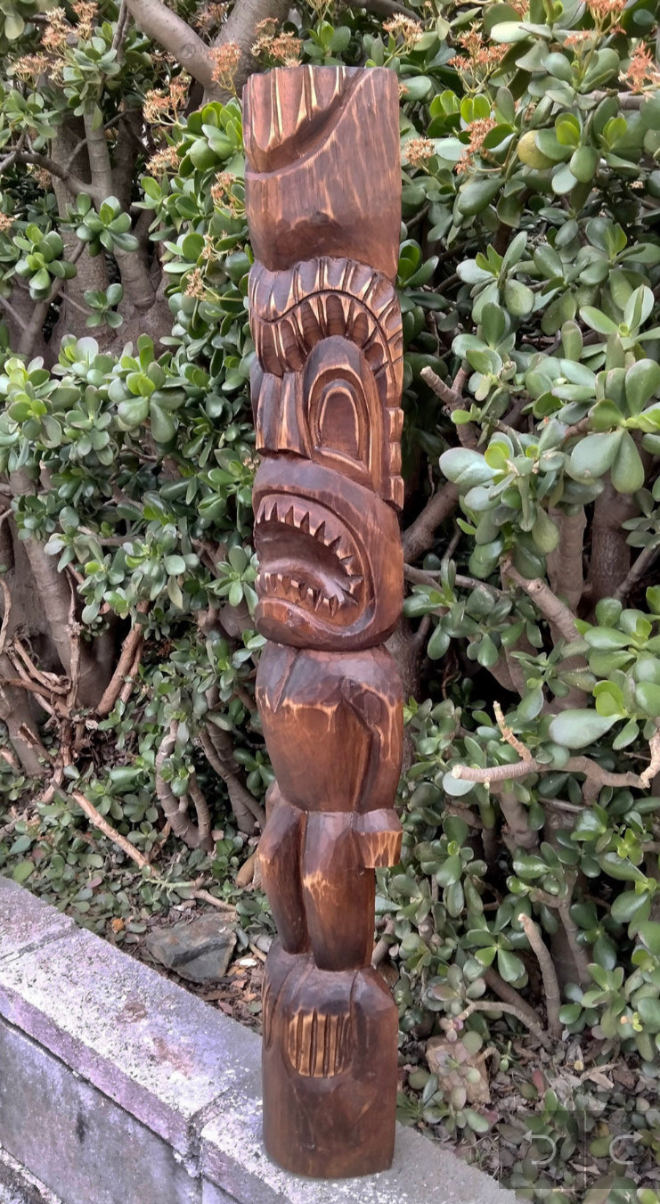 Intricately carved wooden tiki totem statue outdoors among green shrubs.