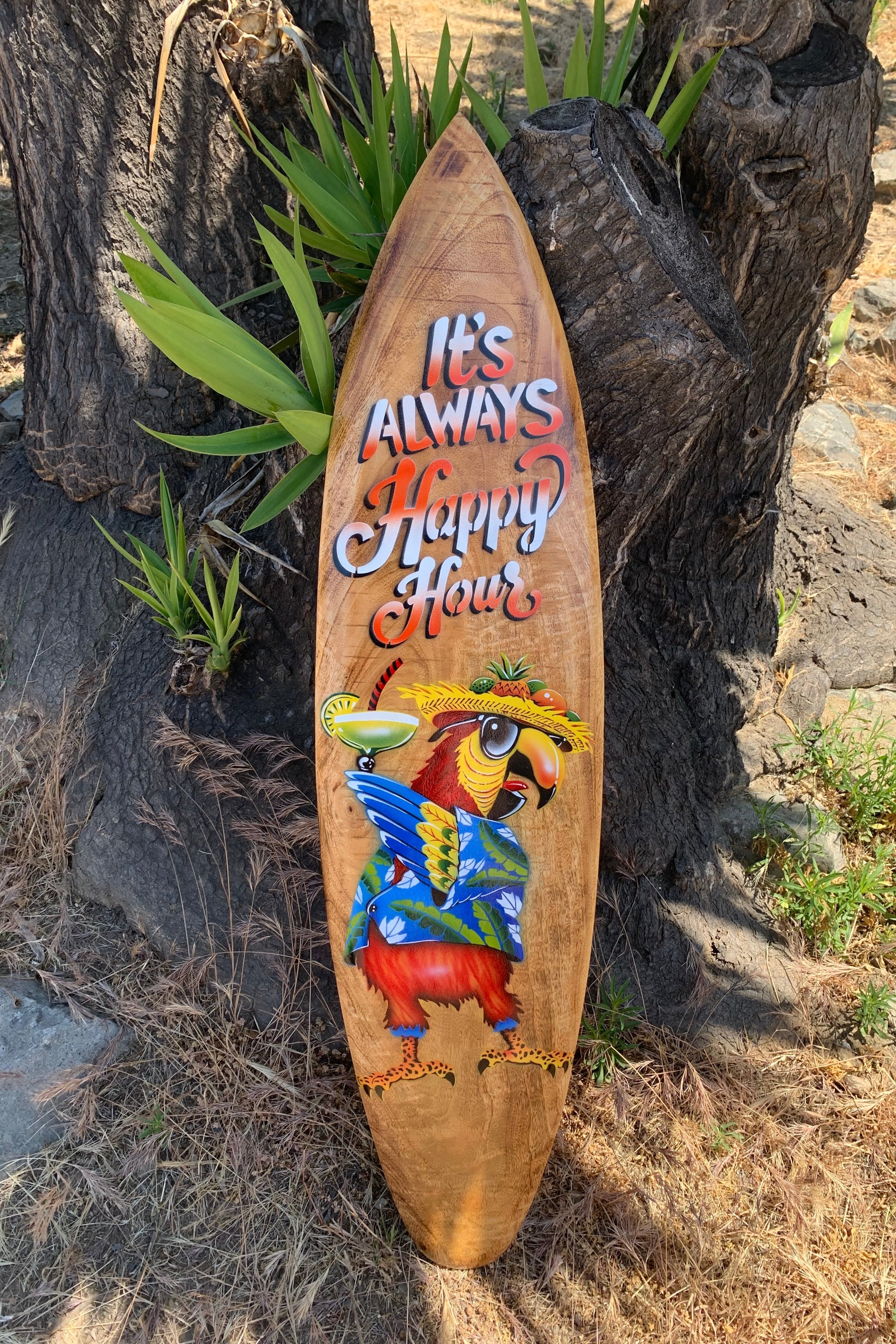 Wooden surfboard with colorful parrot wearing a hat, holding a cocktail, beside a tree; reads 'It's Always Happy Hour'.