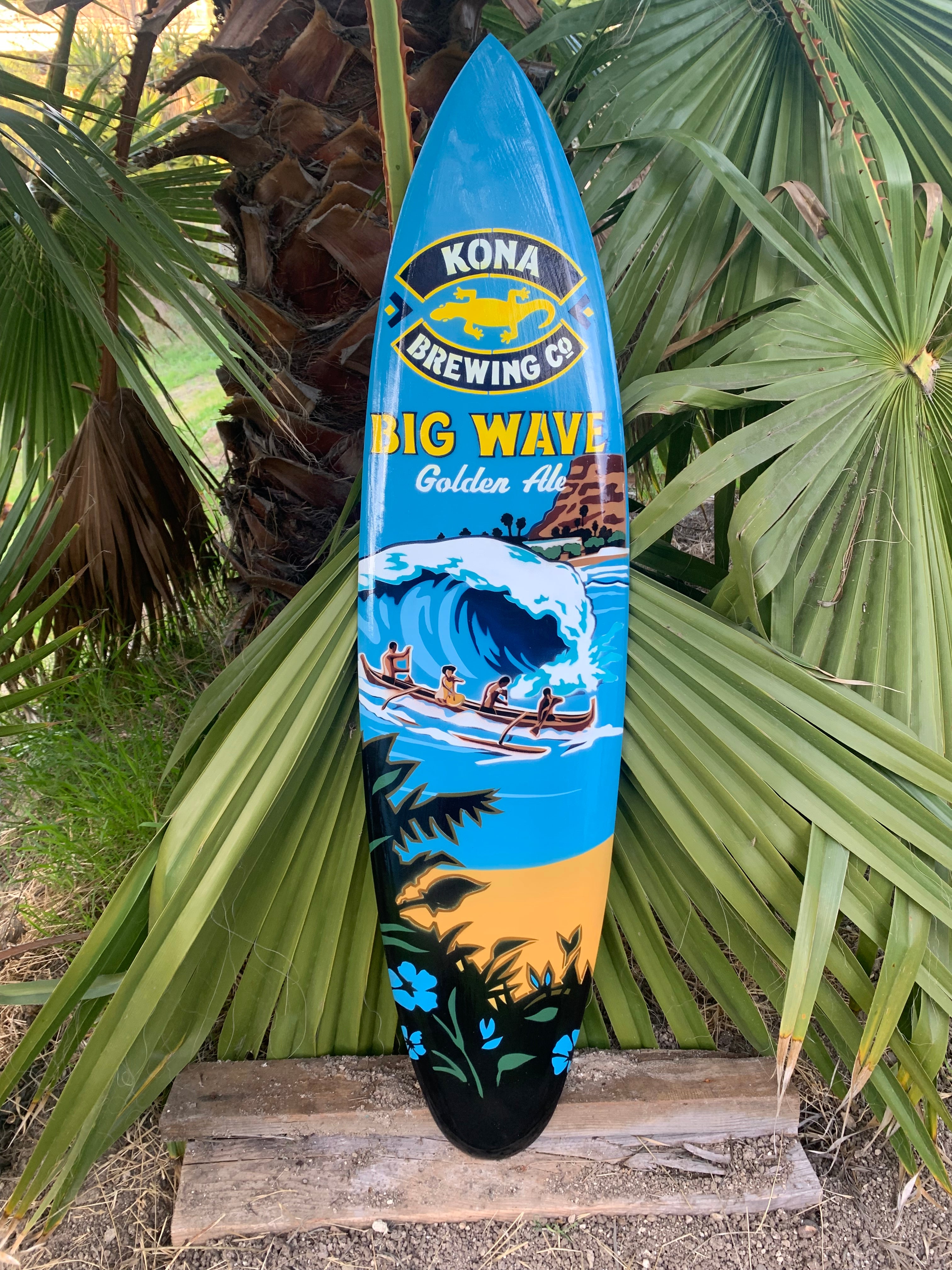 Blue surfboard featuring Kona Big Wave Golden Ale artwork leaning against palm fronds