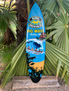 Blue surfboard featuring Kona Big Wave Golden Ale artwork leaning against palm fronds