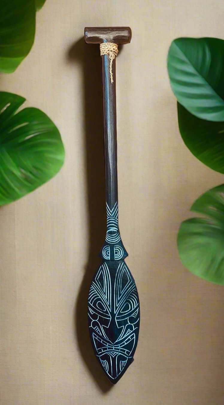 Dark wooden paddle with white tribal geometric patterns and a rope-bound handle, surrounded by green leaves
