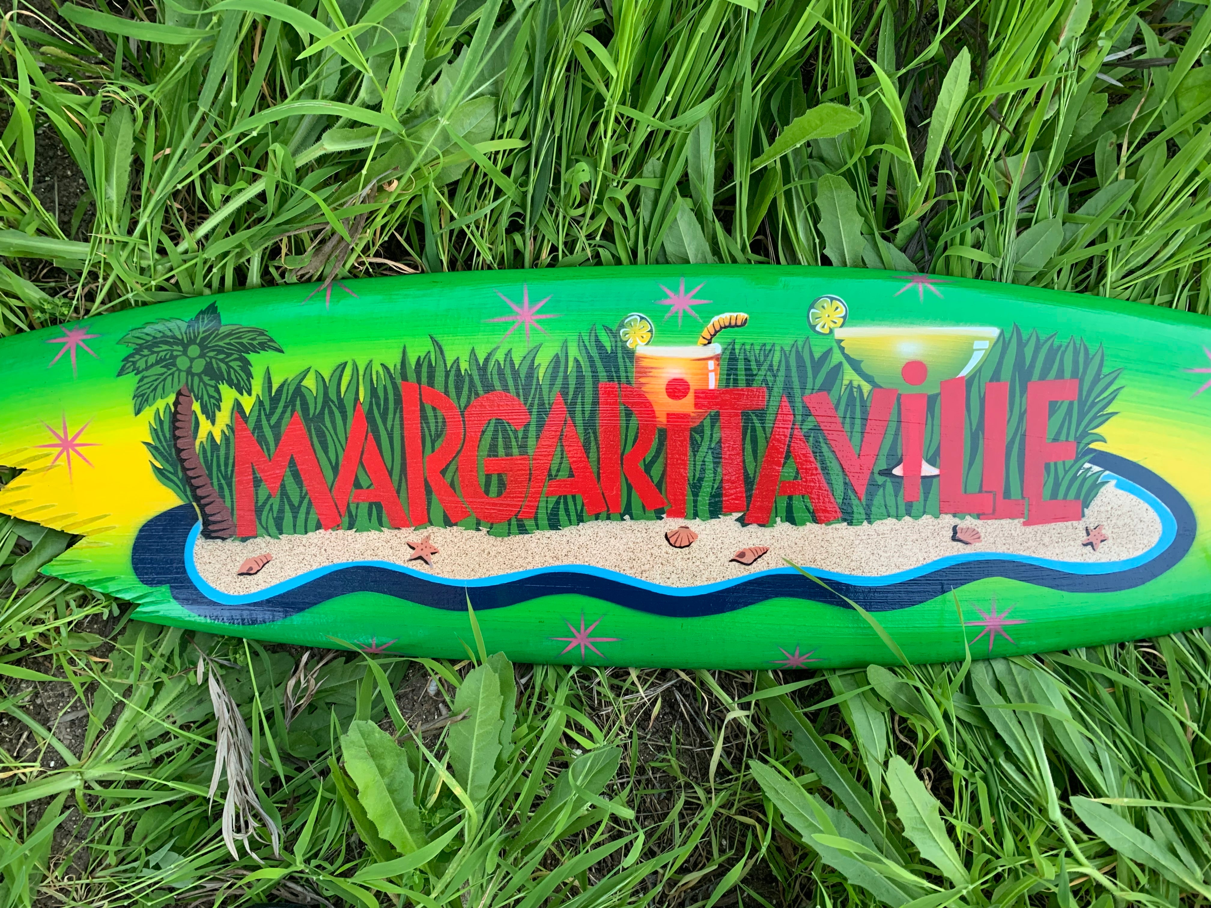 Colorful Margaritaville surfboard on grass with tropical beach scene and palms.