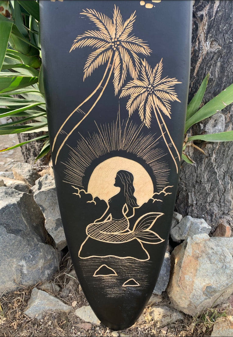 Gold-engraved mermaid on a rock with palm trees and sun on a black surfboard