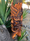 Orange and black carved wooden mask with elaborate details mounted on a tree, surrounded by green leaves.