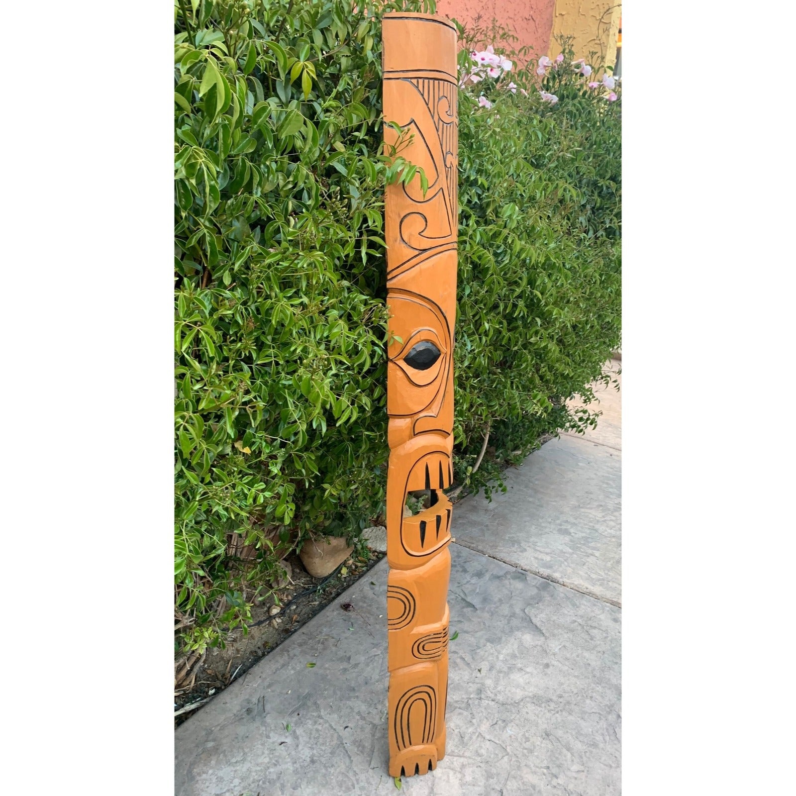 orange carved tiki totem pole with abstract patterns beside a green hedge outdoors