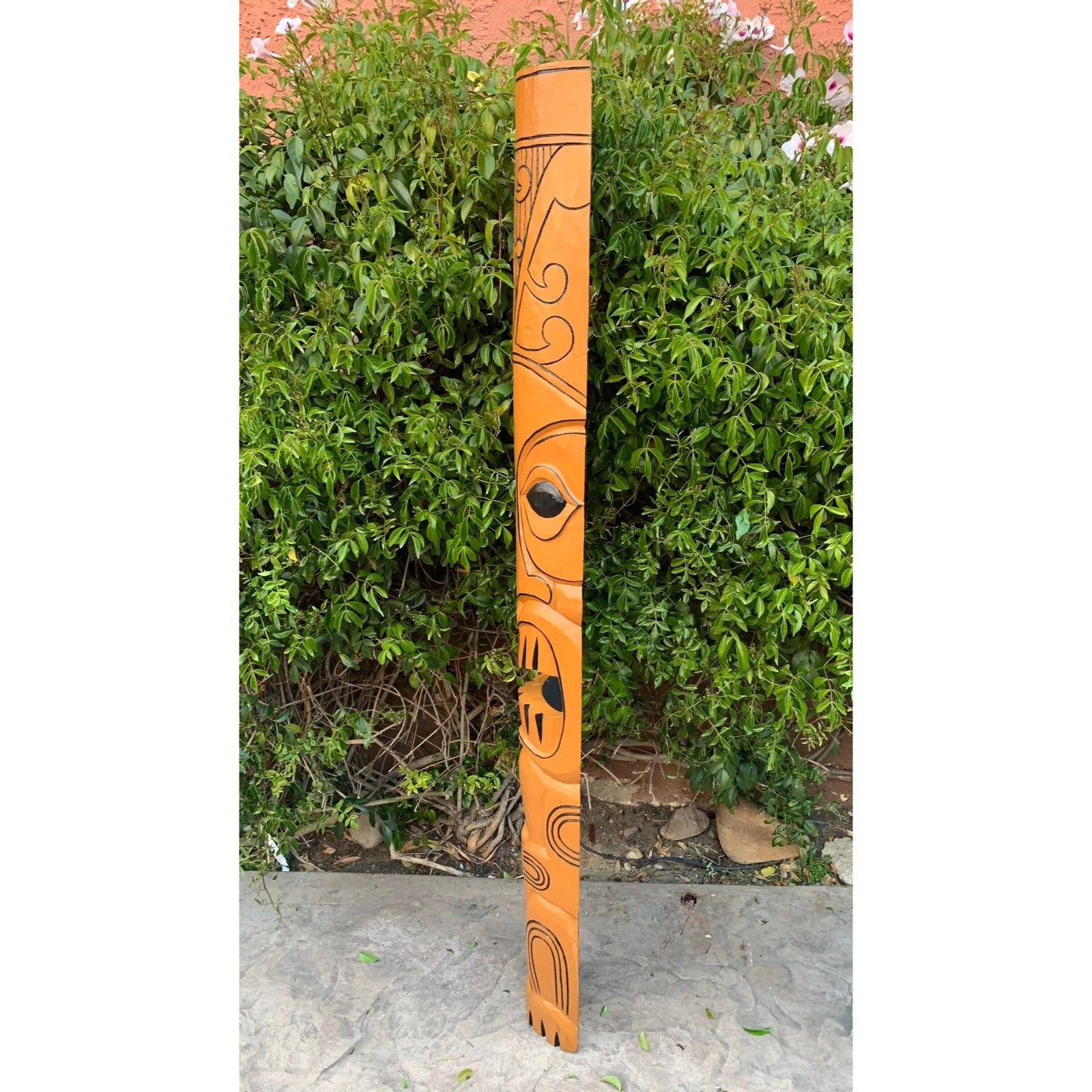 Orange totem pole with black abstract carvings beside a green hedge