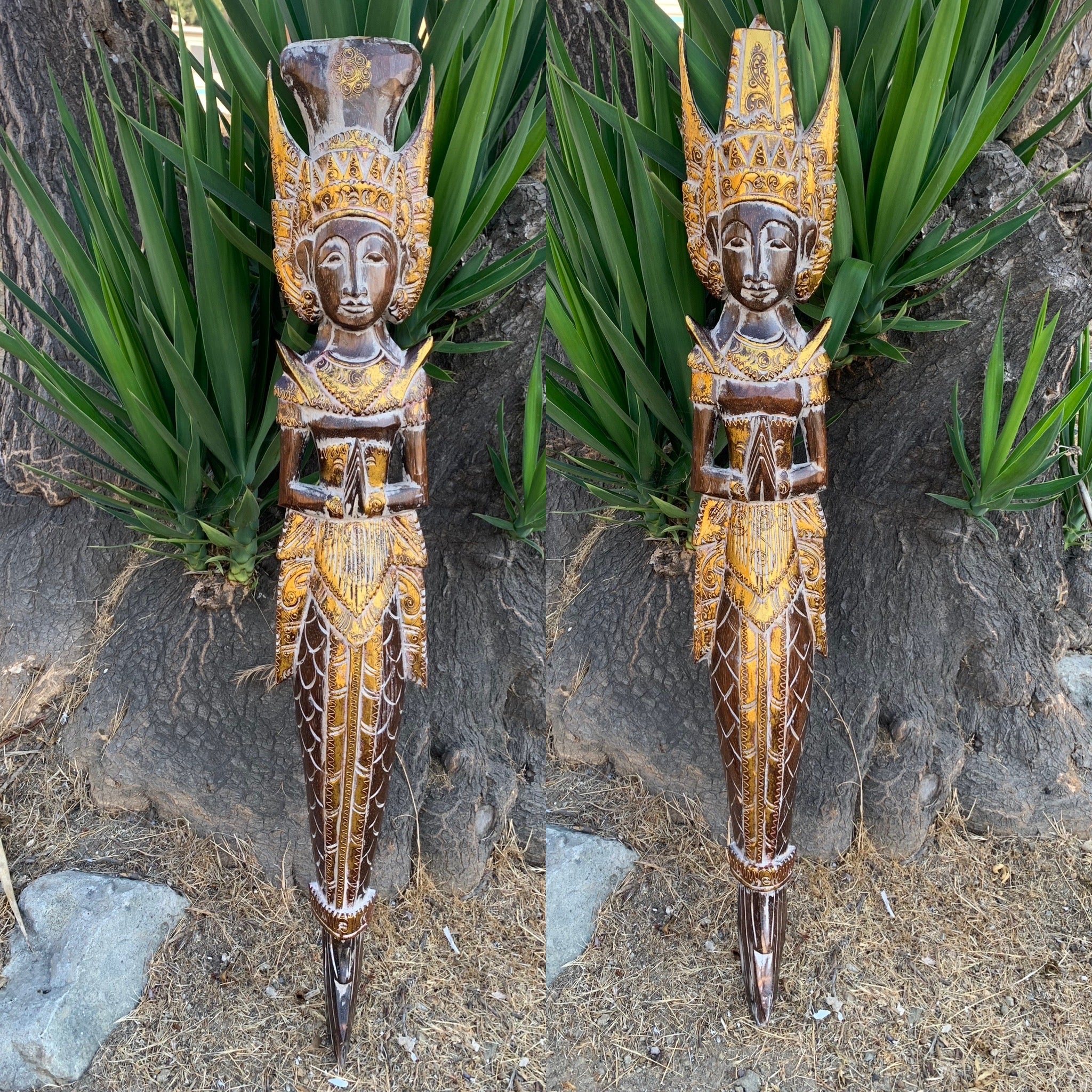 Two ornate carved wooden statues with gold accents and tall headdresses outdoors among plants