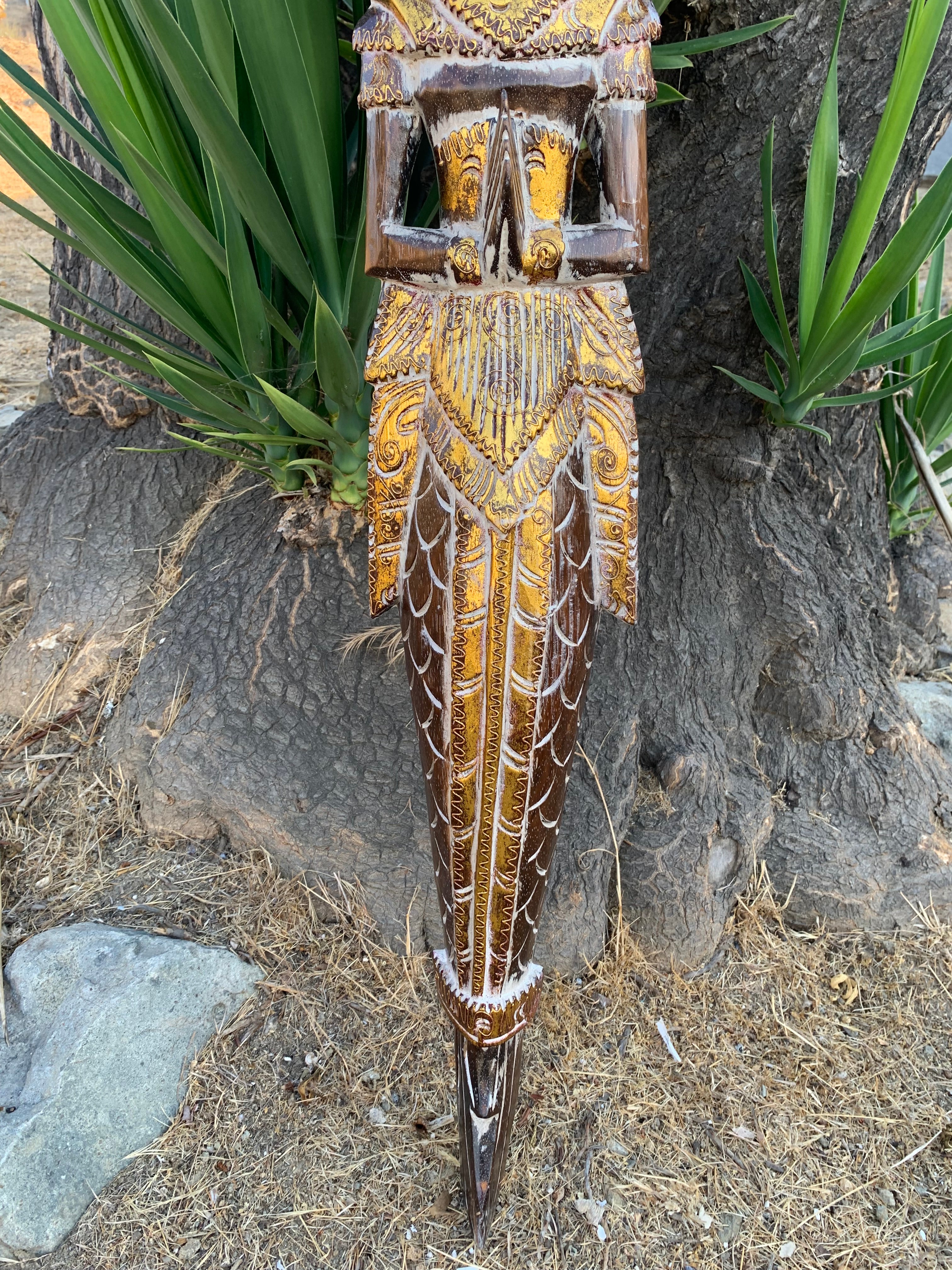 Ornate carved wooden tiki ceremonial staff with gold detailing, hanging by a tree.