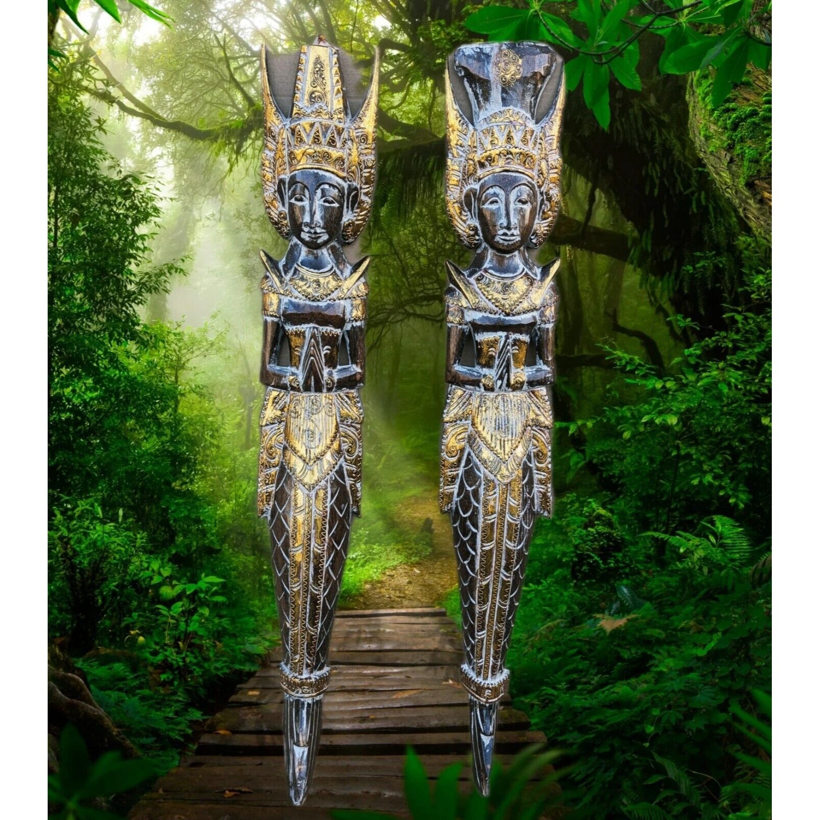 Two tall ornate carved wooden statues with gold detailing standing on a wooden path in a lush jungle.