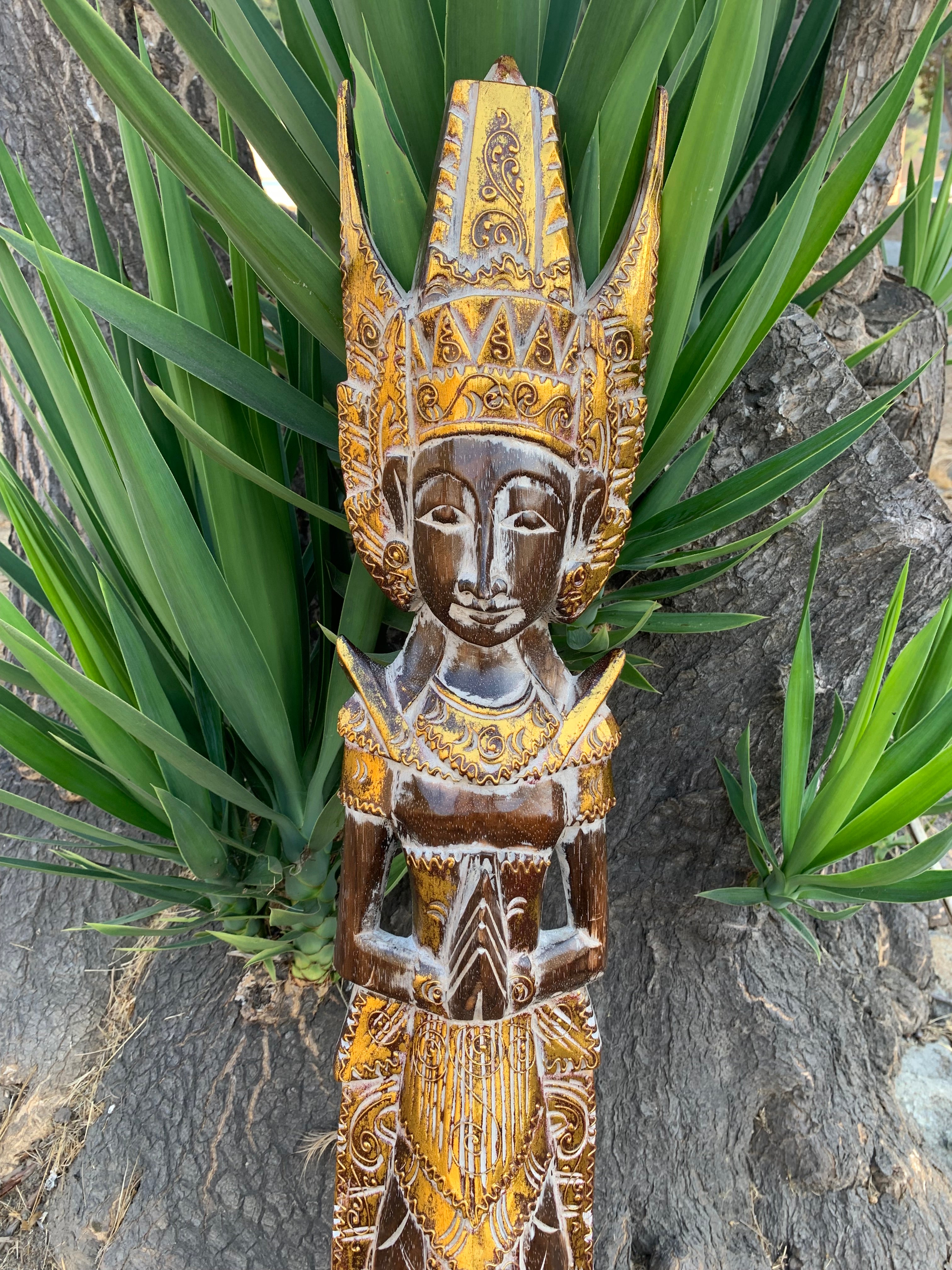 Carved wooden statue with ornate gold detailing and tall headdress, set among green plants.